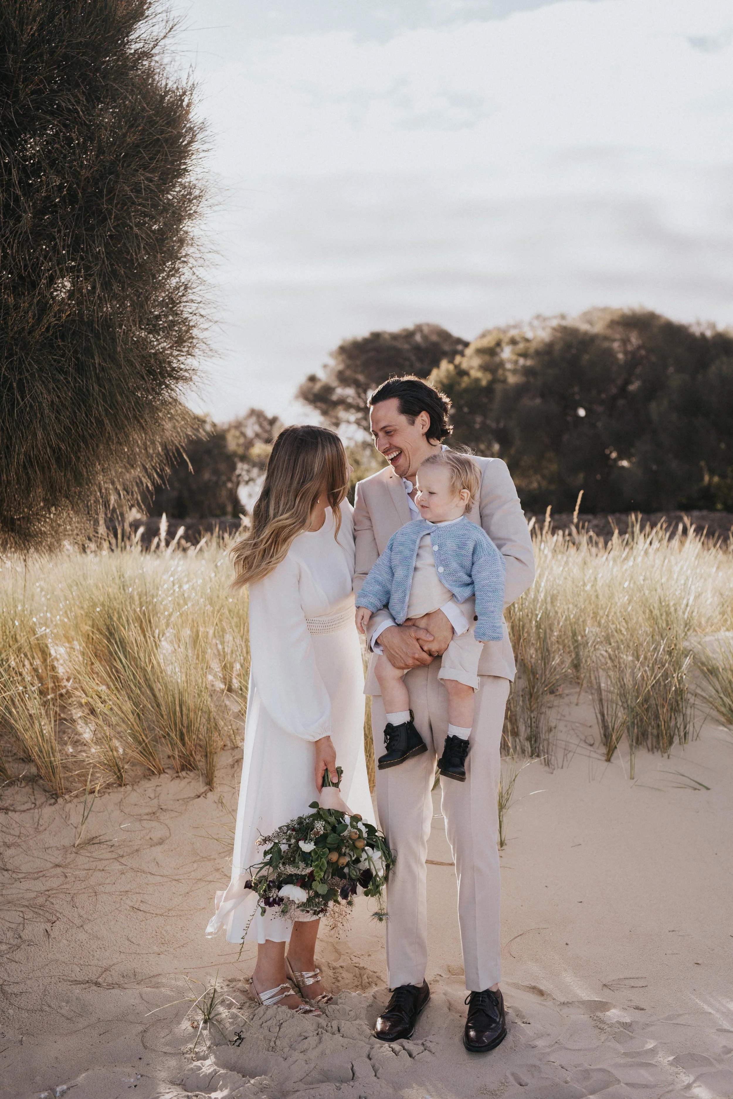 A happy family standing on a sandy beach with tall grass and trees in the background, during a wedding or special occasion.