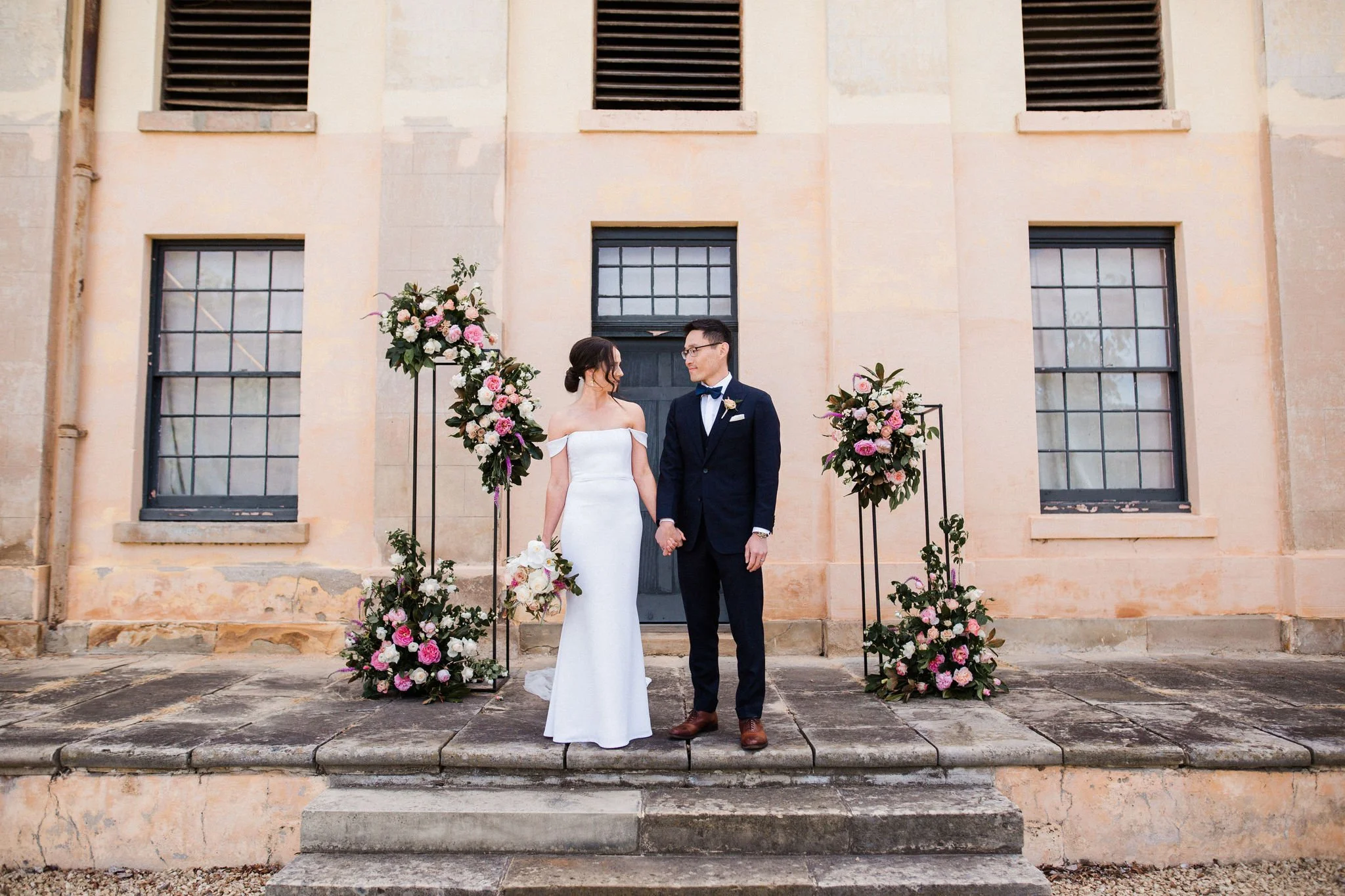 Bride and groom holding hands on wedding altar decorated with floral arrangements outside a historic building.