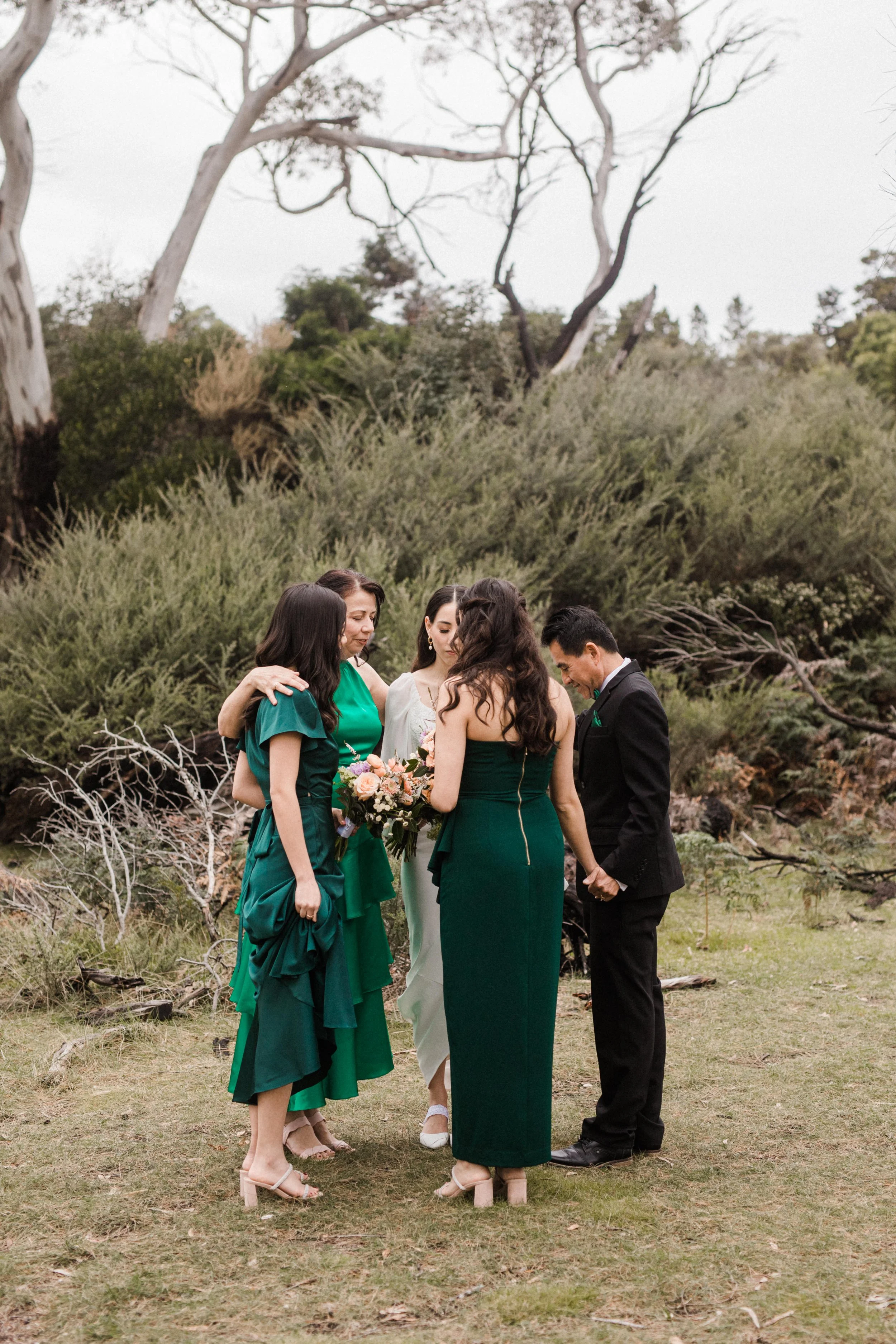 A group of four women and one man standing outdoors surrounded by trees, with the women holding a bouquet of flowers, appearing to participate in a wedding ceremony.