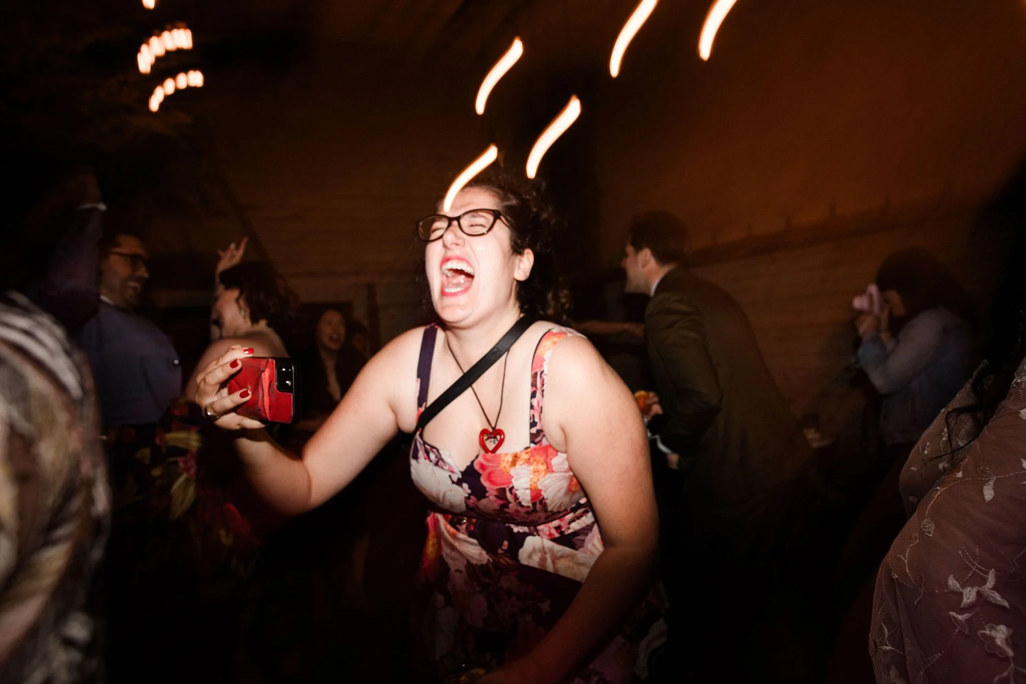 A woman with glasses and curly hair wearing a floral dress, singing or shouting with joy at a party or celebration, holding a red phone in her hand, with other people dancing and talking in the background.