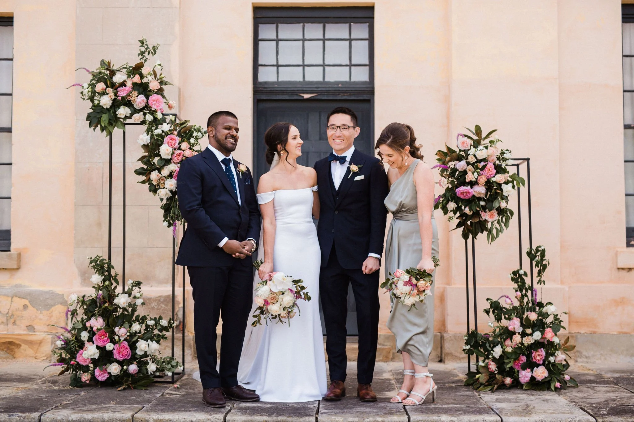 A wedding ceremony with five people standing outdoors in front of a building with a black door and large floral arrangements. The bride in a white off-shoulder gown and the groom in a dark suit with a bow tie are in the center, smiling and looking at