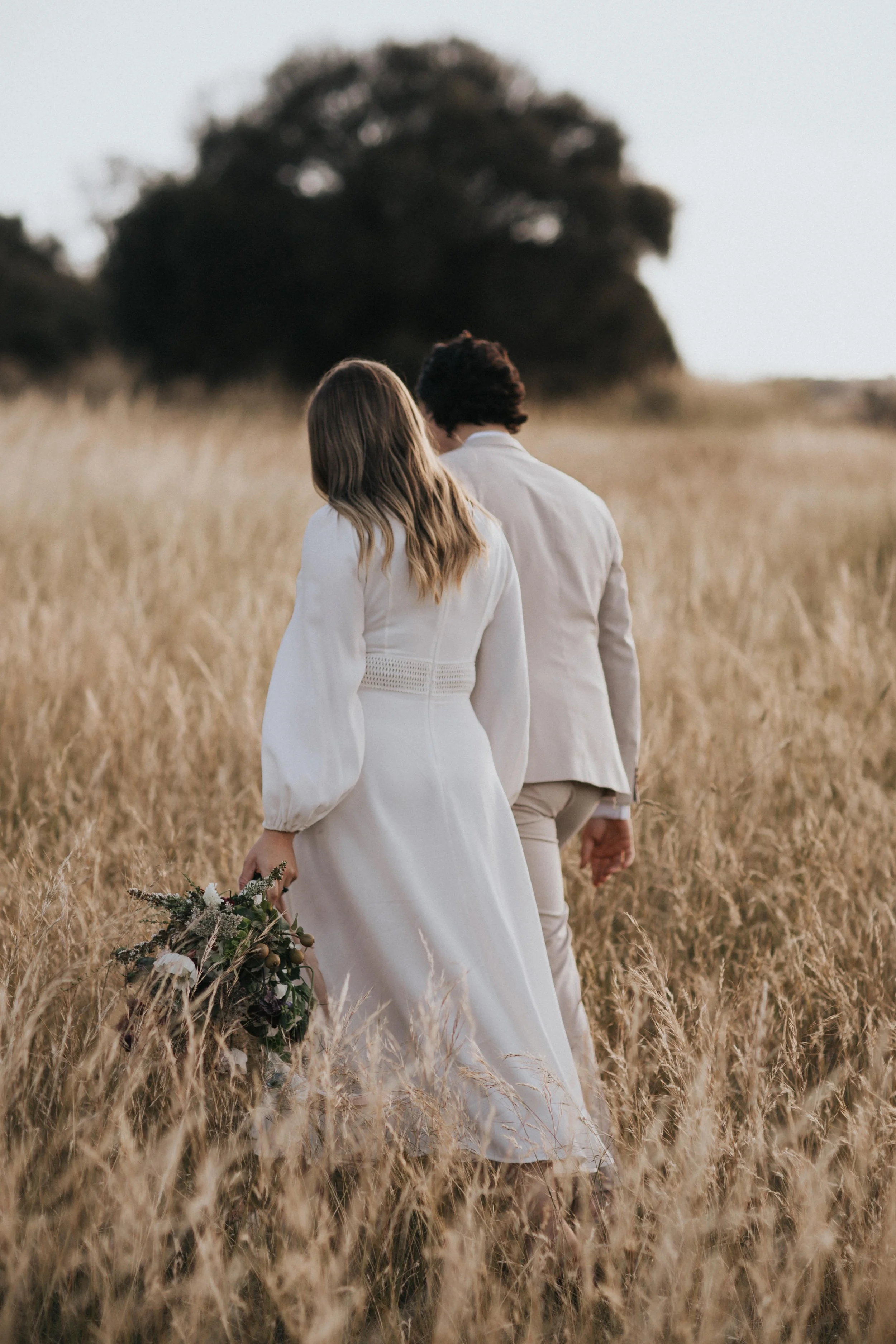 A couple dressed in light-colored wedding attire walking in a field of tall golden grass, with one holding a bouquet of flowers and their backs facing the camera.