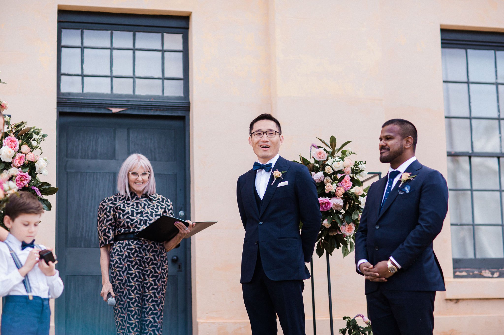 A wedding ceremony outdoors with a bride and groom standing in front of a building with large windows, accompanied by a woman officiant and a young boy with a camera. The bride and groom are dressed in formal suits and are smiling.