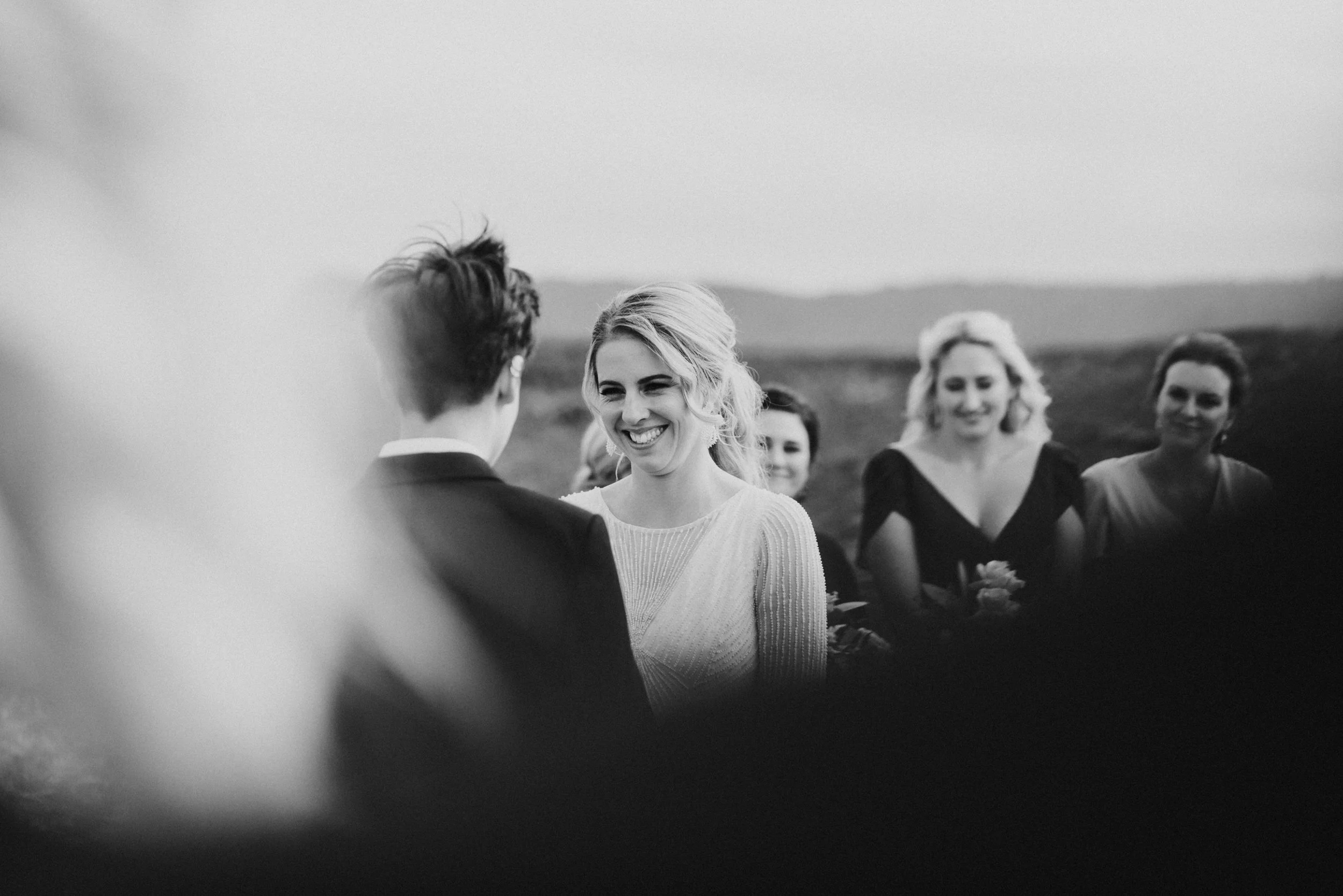 Black and white photo of a smiling woman with blonde hair, dressed in a light-colored dress, outdoors with a group of women in the background, mountains in the distance.