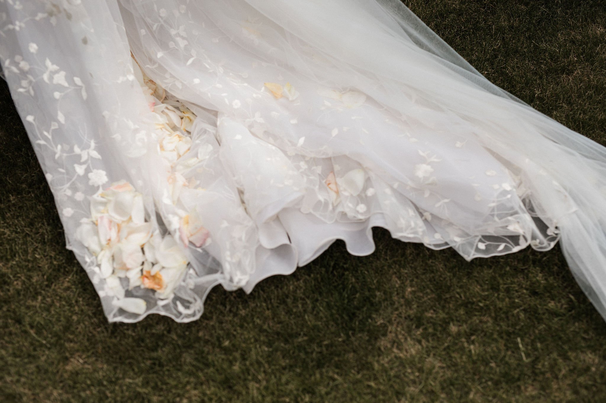 A wedding dress with floral lace details lying on the grass, with rose petals scattered at the bottom of the dress.
