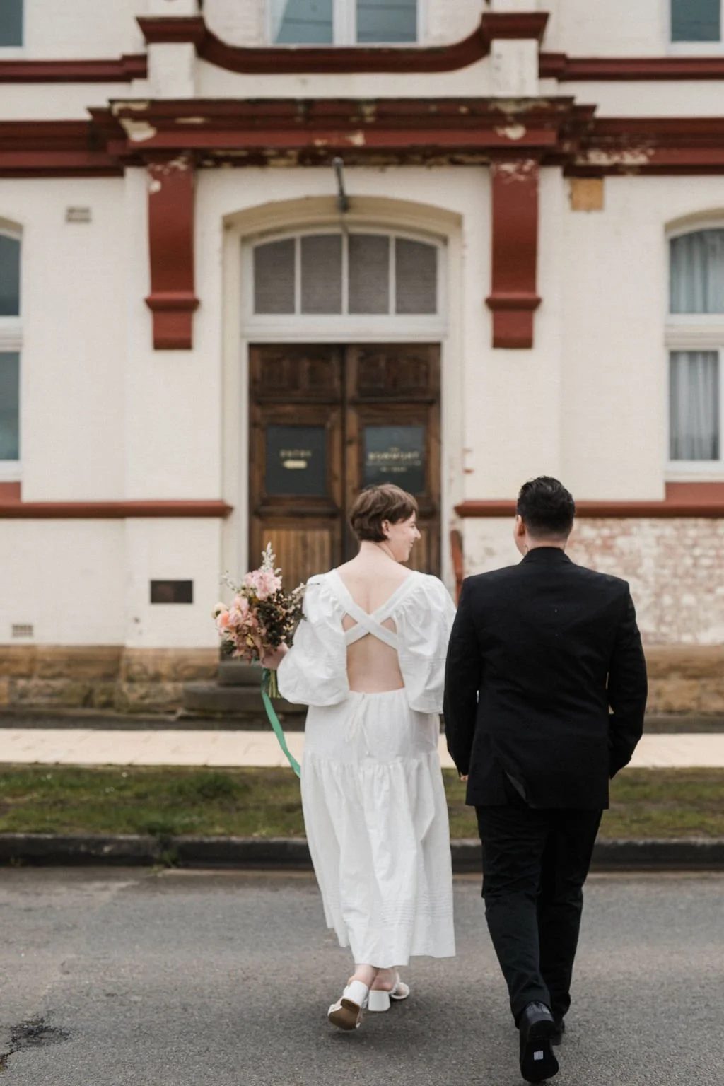 A couple, with the woman holding a bouquet of flowers, walks hand-in-hand in front of an old building with peeling paint and boarded-up windows.