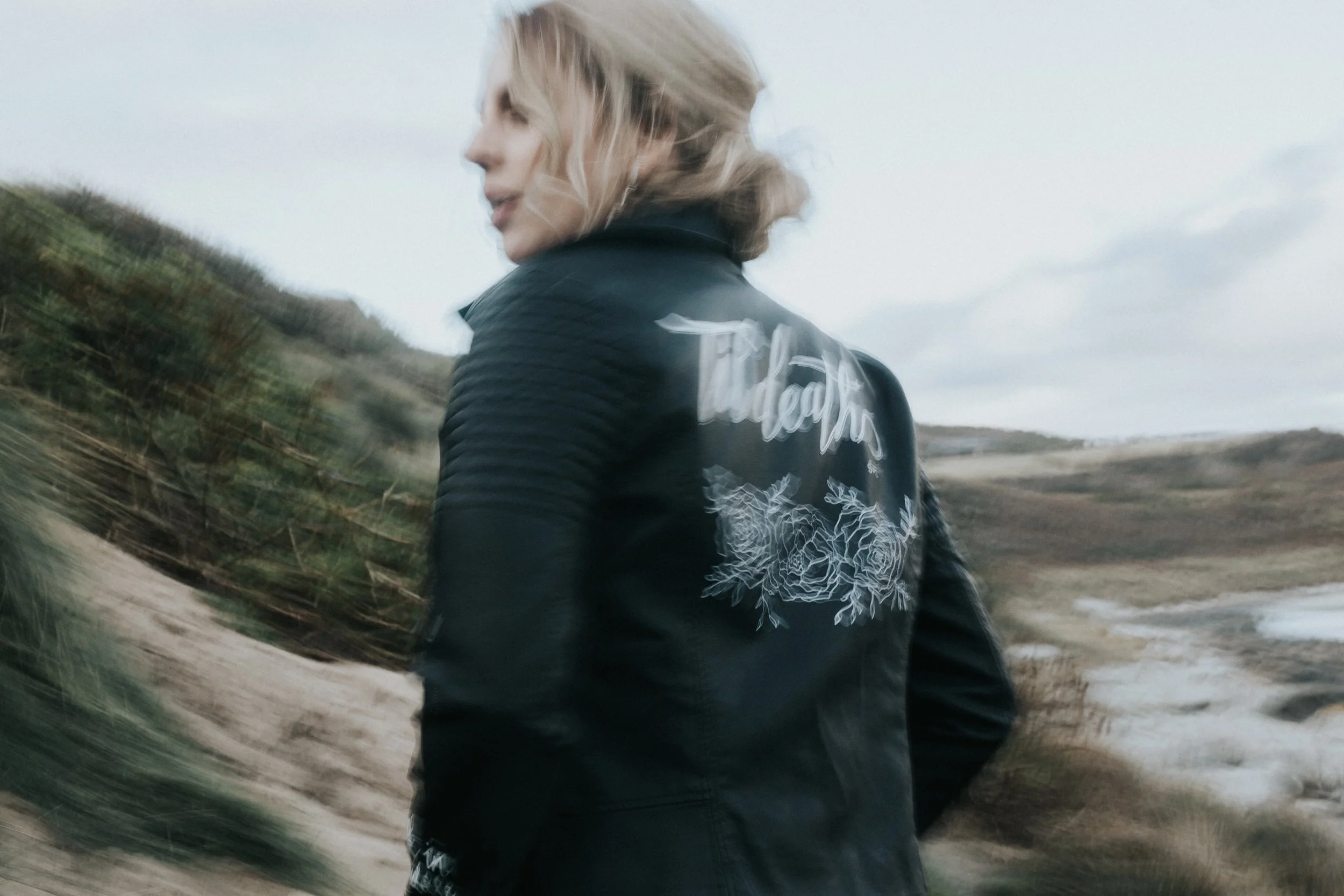 A woman with blonde hair wearing a black jacket with the word "Tidepools" and floral design on the back, standing outdoors near a coastal landscape with hills and water.