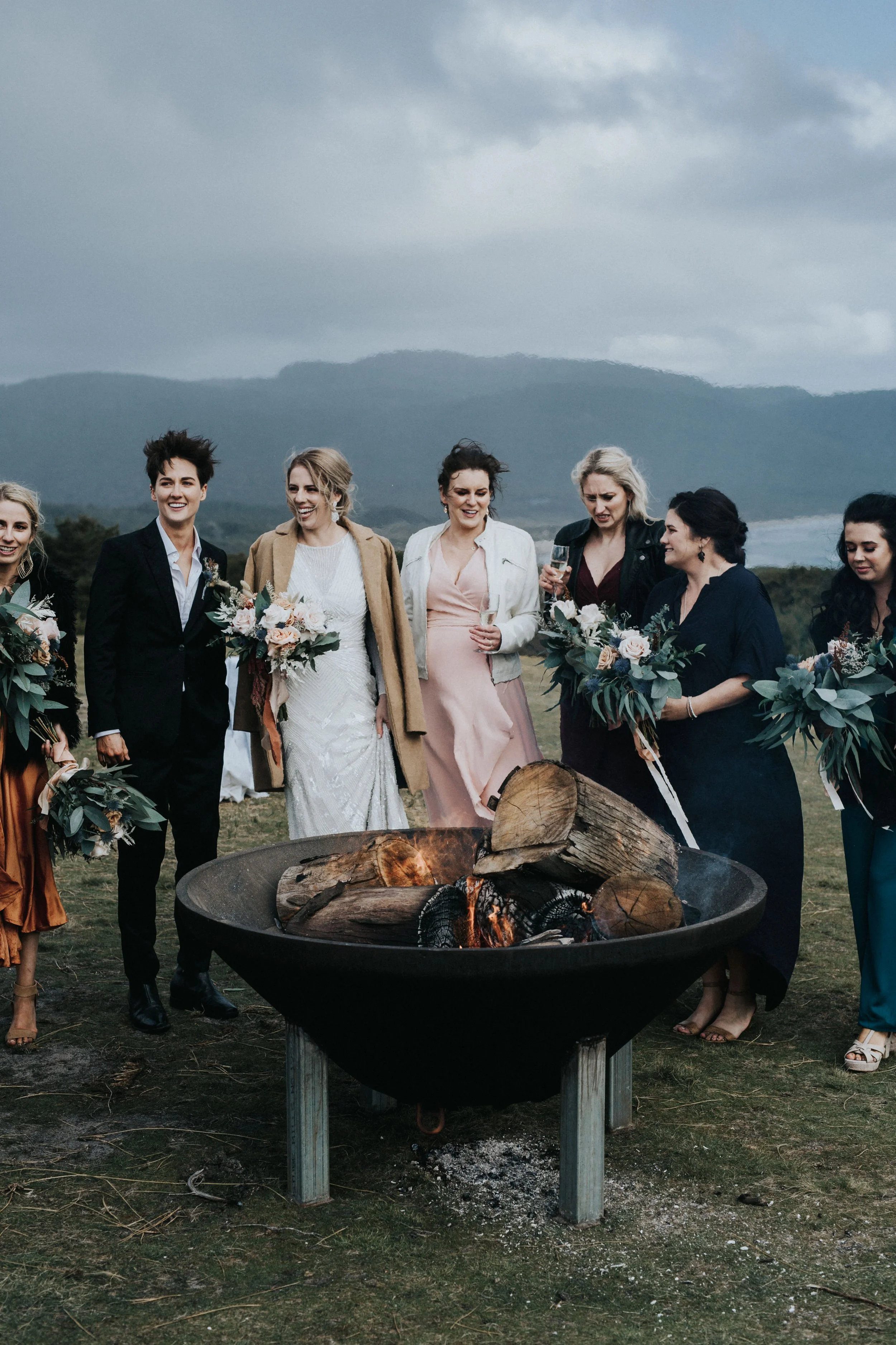Group of people gathered around a fire pit outdoors, at a wedding, with mountains and cloudy sky in the background.