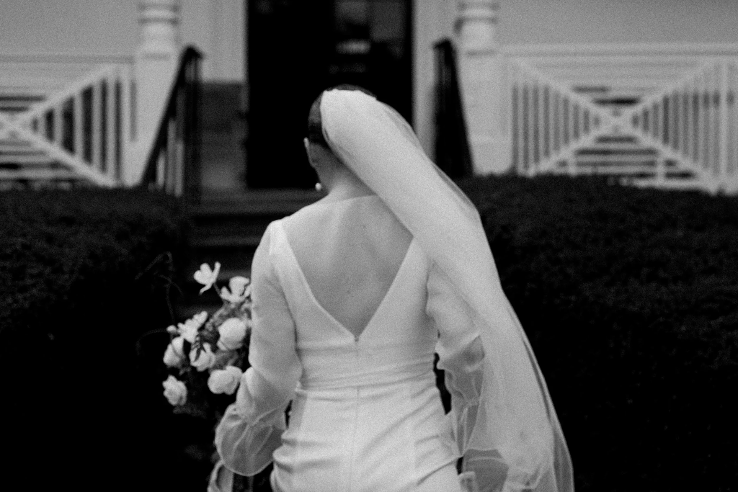 A bride in a wedding dress with a long veil walking away on a path towards a house or building with stairs and a porch.