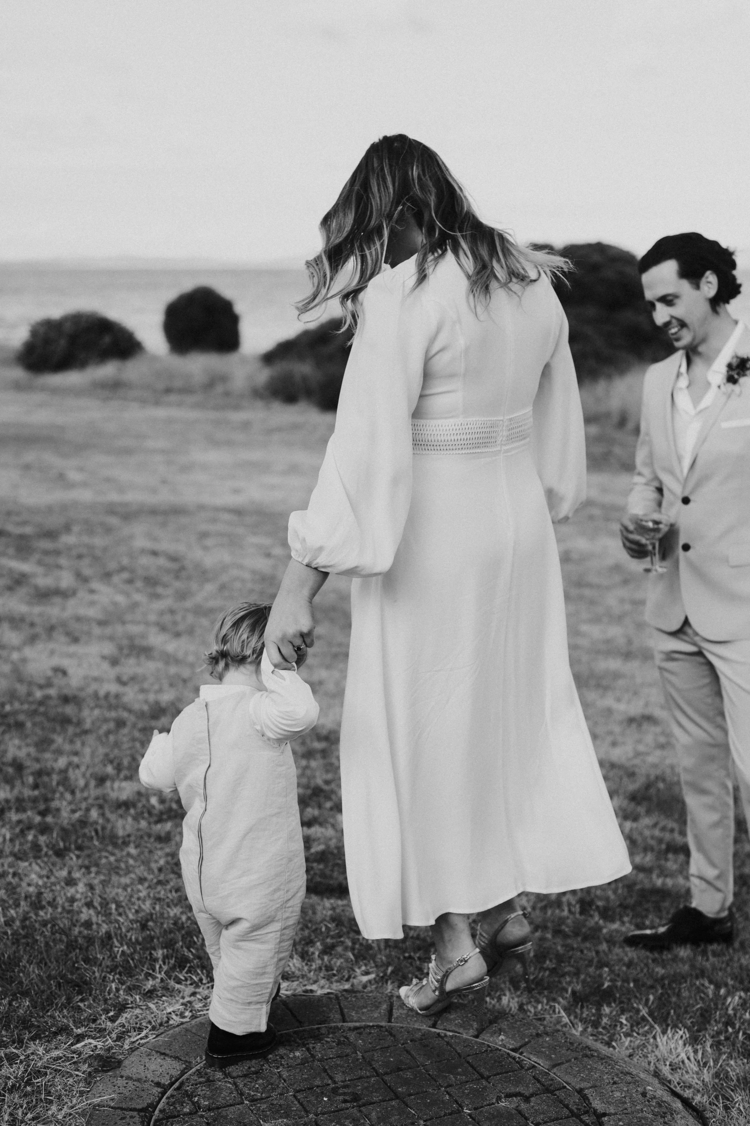 A woman dressed in a long dress holding a young child's hand, standing outdoors on a grassy area, with a man in a suit nearby, smiling in the background.