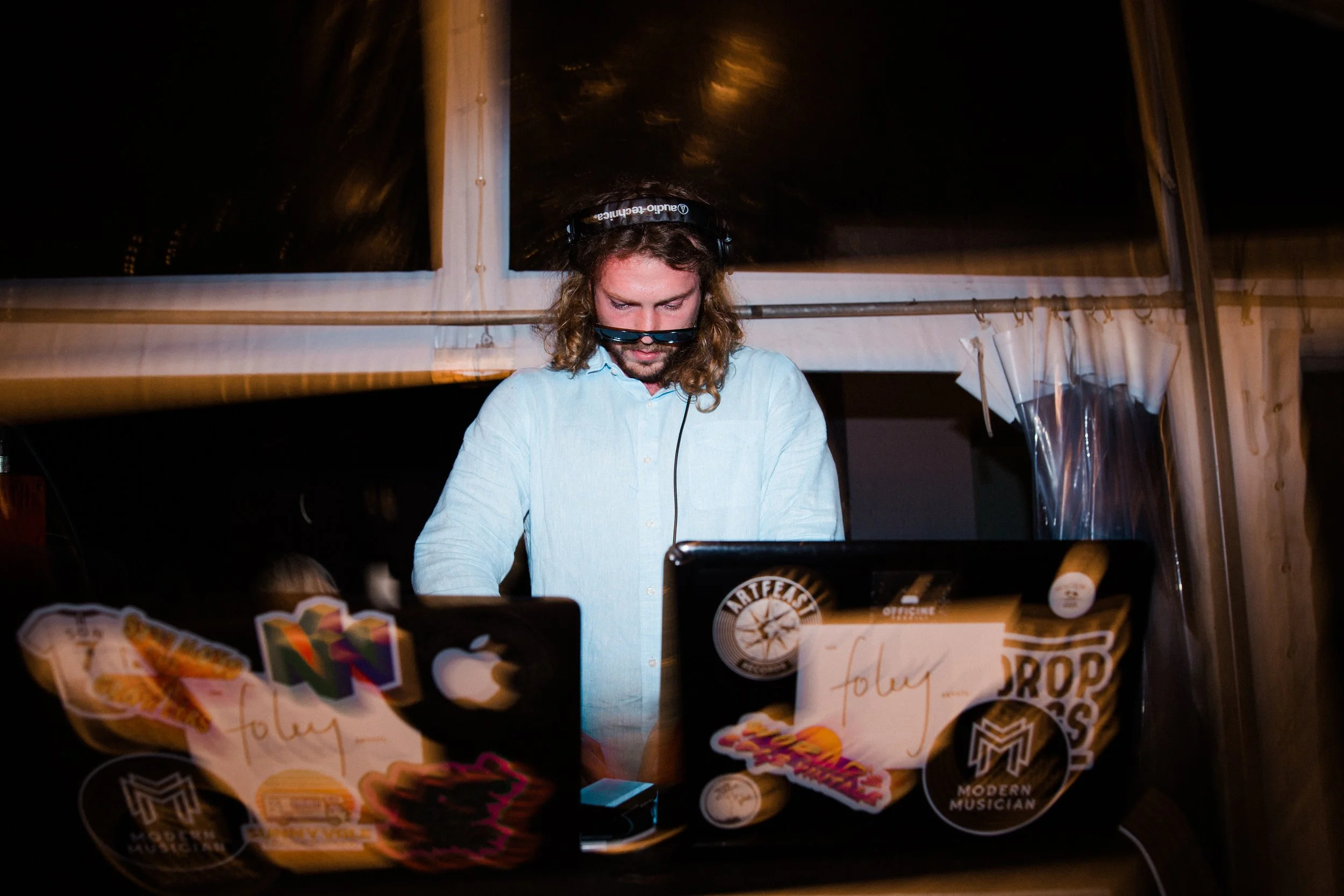 A DJ with long curly hair wearing headphones and sunglasses, working at a laptop covered in stickers, in a tent or enclosed space at night.