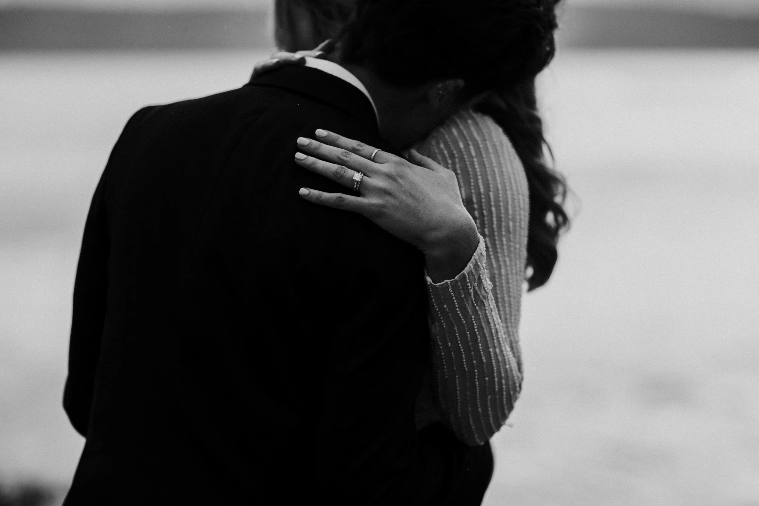 Black and white photo of a couple embracing, with the woman's hand showing an engagement ring resting on her partners shoulder.