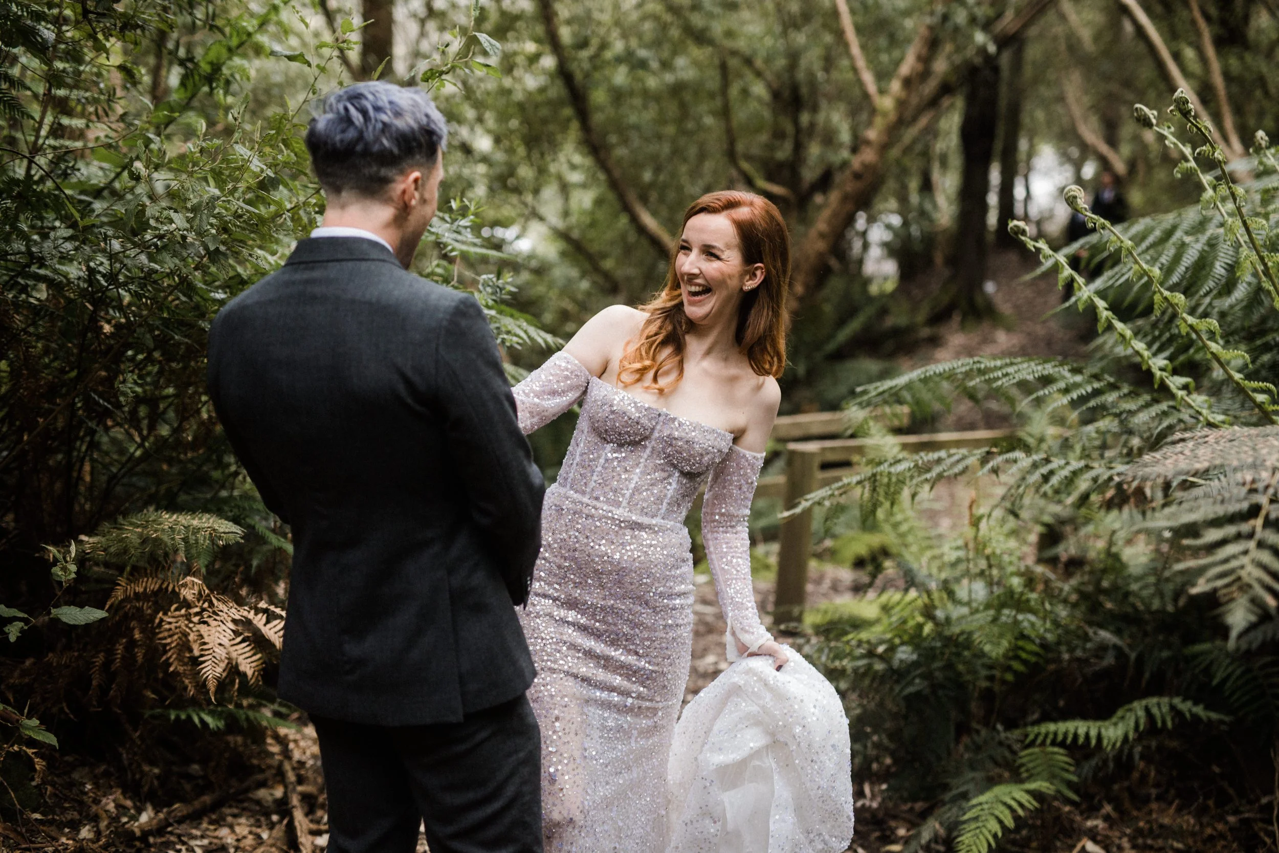 A woman in a sparkly off-the-shoulder dress laughing and holding her dress, standing outdoors with a man in a dark suit in a wooded area.