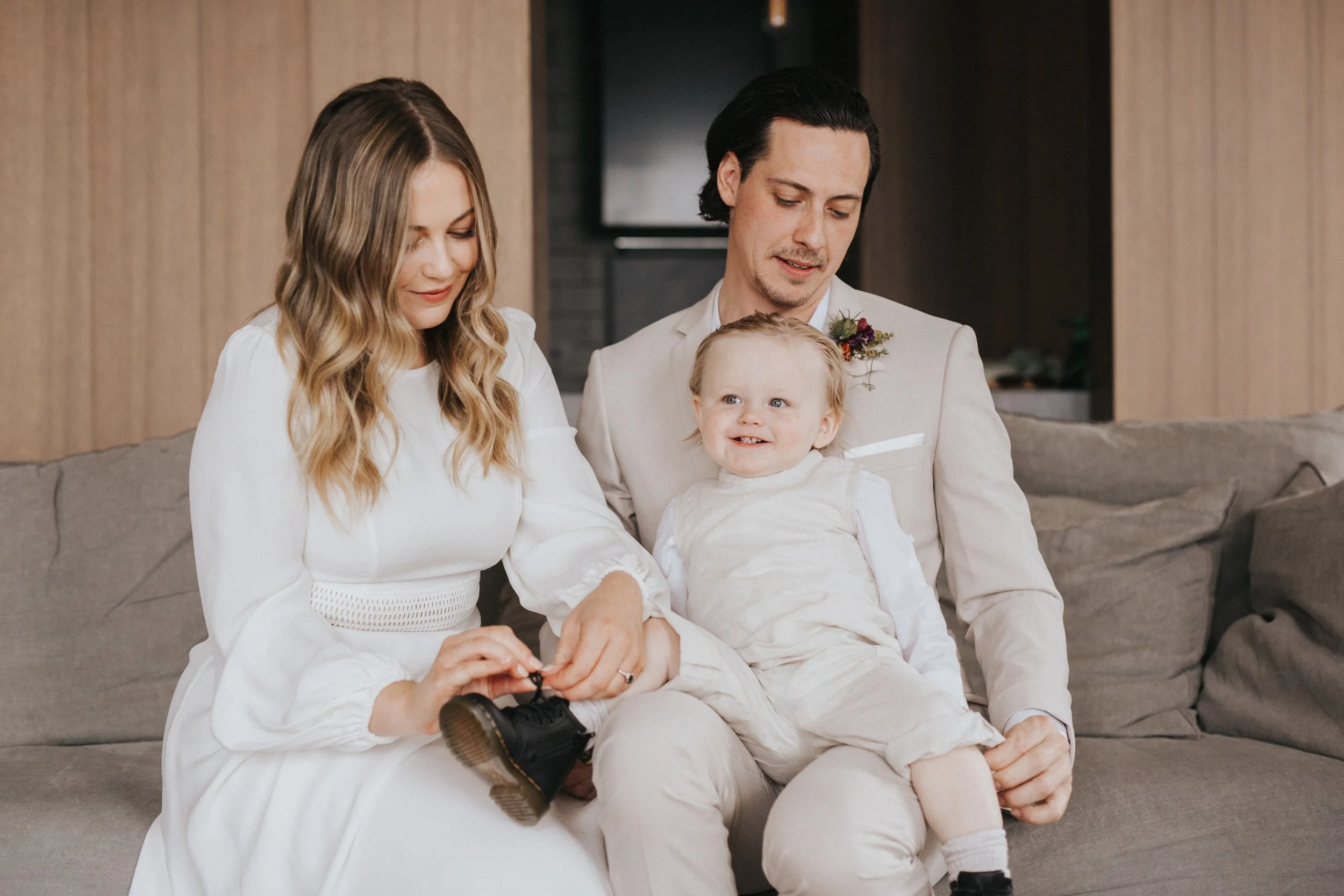A family of three sitting on a sofa, with a woman tying the shoelaces of a young boy, while a man looks on. They are indoors, dressed in light-colored clothes, smiling and engaging with each other.