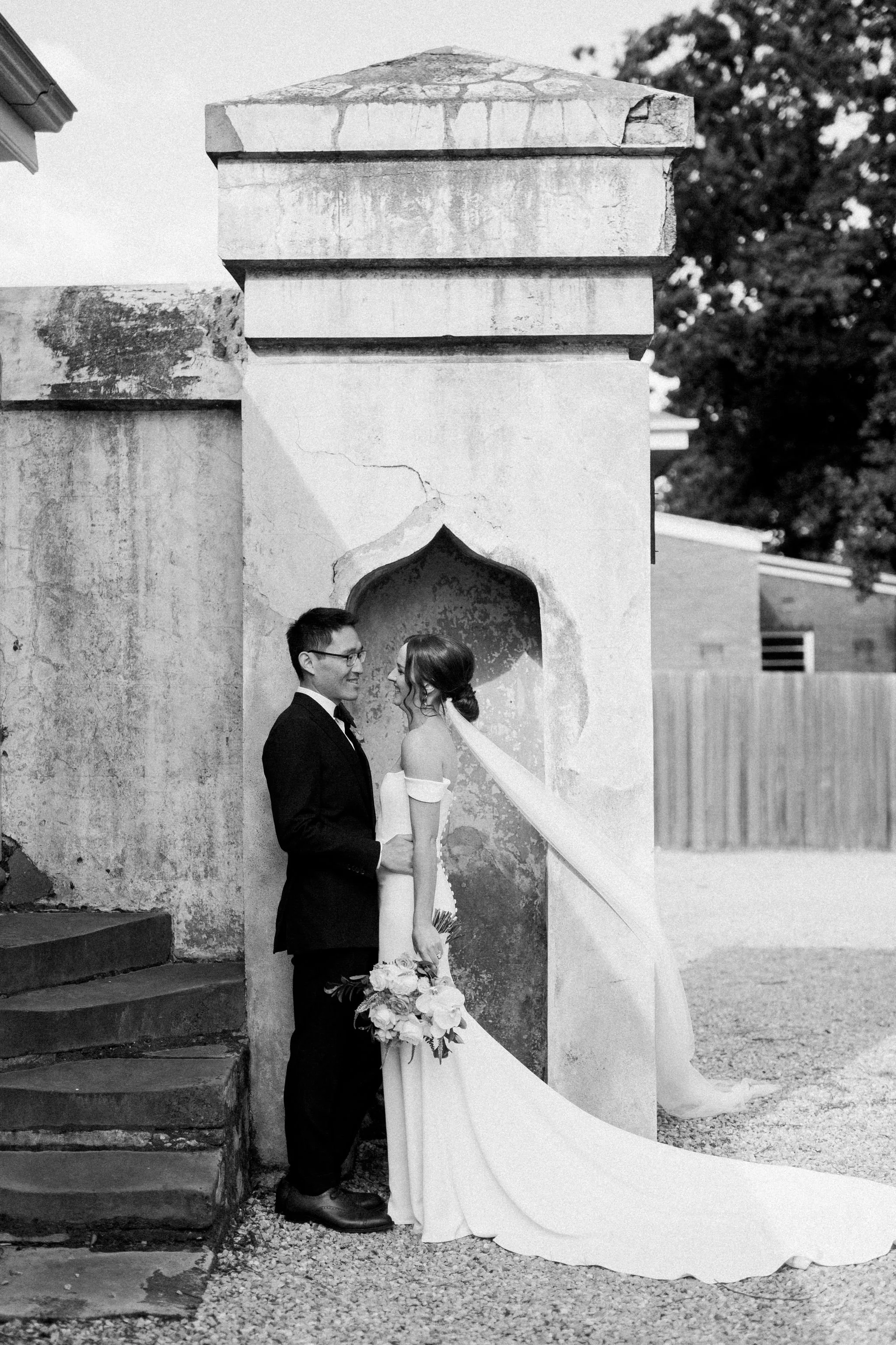 A newlywed couple stands close to each other, smiling, in front of an aged, textured wall with a niche. The groom wears a dark suit and glasses, holding a bouquet of flowers, while the bride wears an off-the-shoulder wedding gown with a long train an
