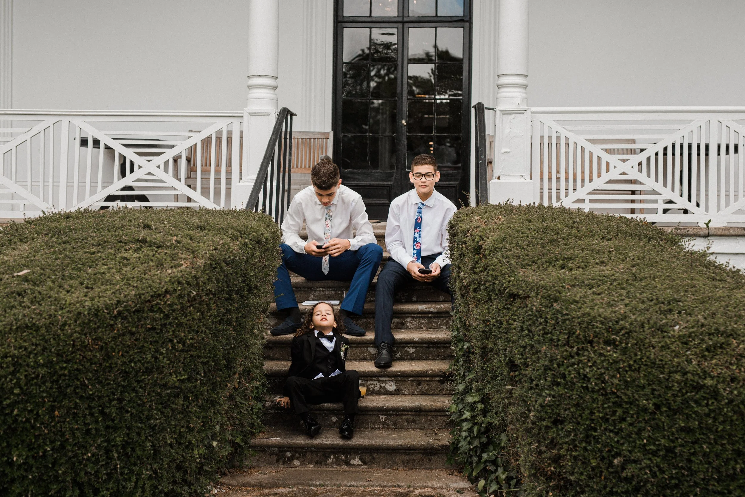 Three boys in formal attire sitting on steps leading to a house, with the youngest lying on the stairs, one older boy looking at a phone, and the other boy looking at his phone with glasses.