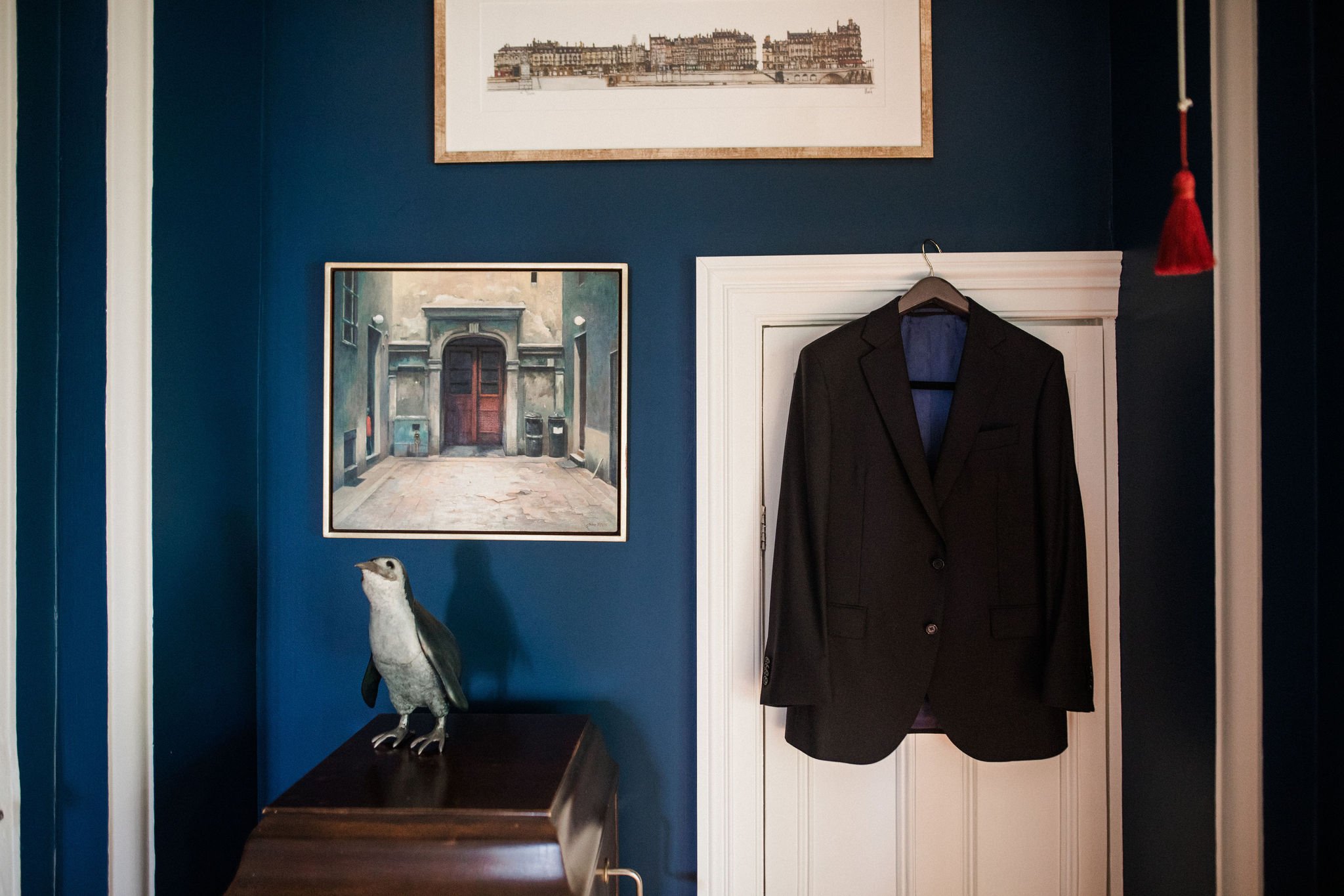 A dark suit jacket hanging on a hook on a white door in a room with dark blue walls. There are two framed pictures on the wall, one of which depicts an outdoor scene with buildings. A penguin figurine is placed on a dark wooden piece of furniture bel