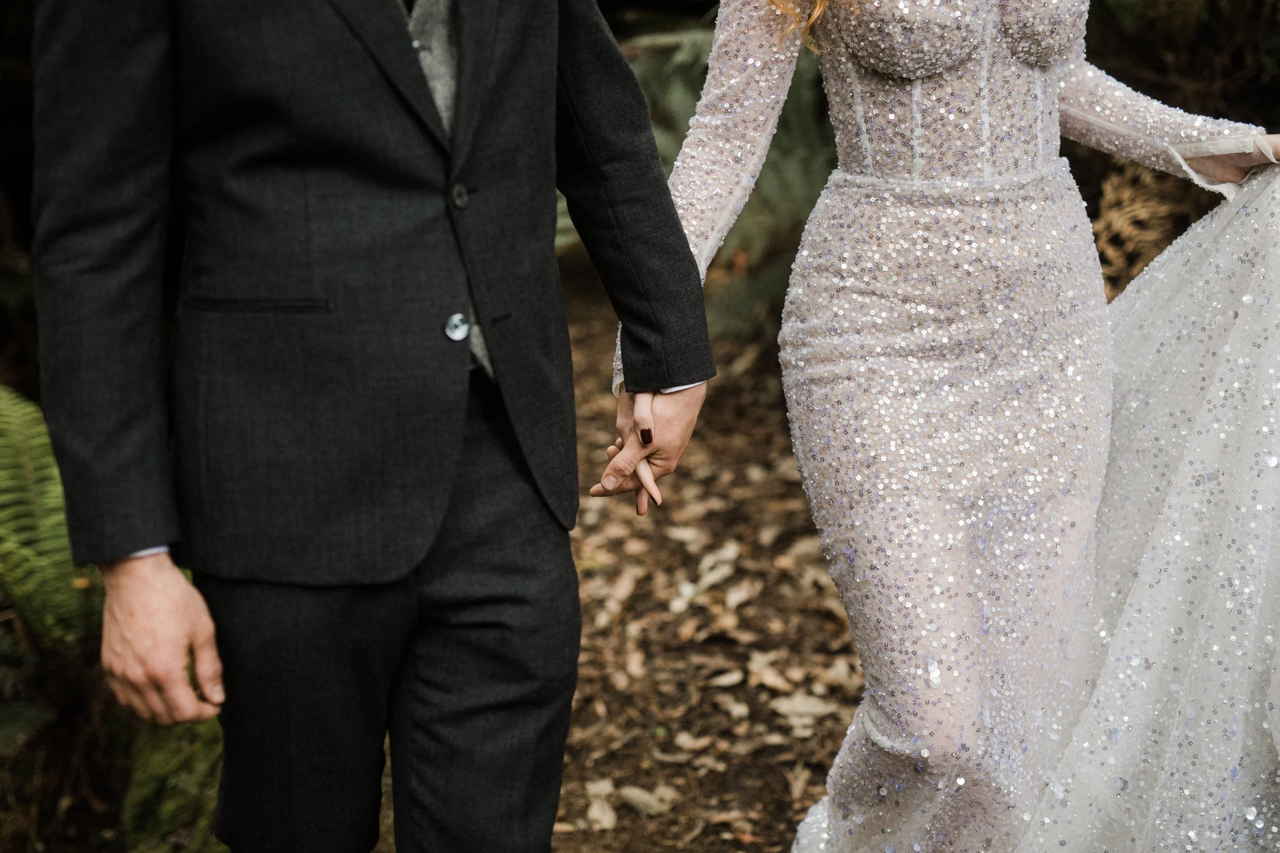 A couple holding hands during a wedding, with the bride wearing a sparkling white gown and the groom in a dark suit, walking outdoors on a leaf-covered ground.