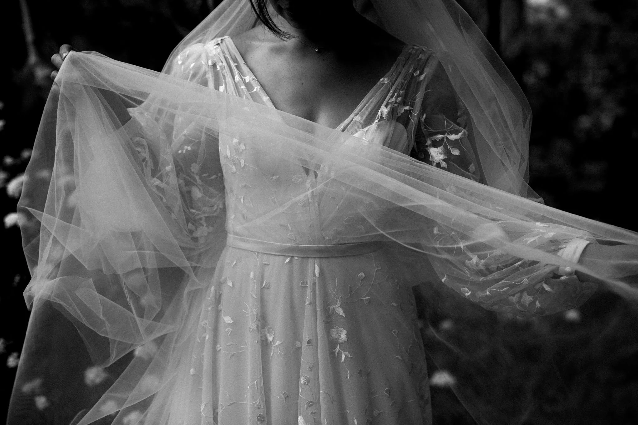 A woman in a delicate, long-sleeved floral dress holding a sheer veil in an outdoor setting.