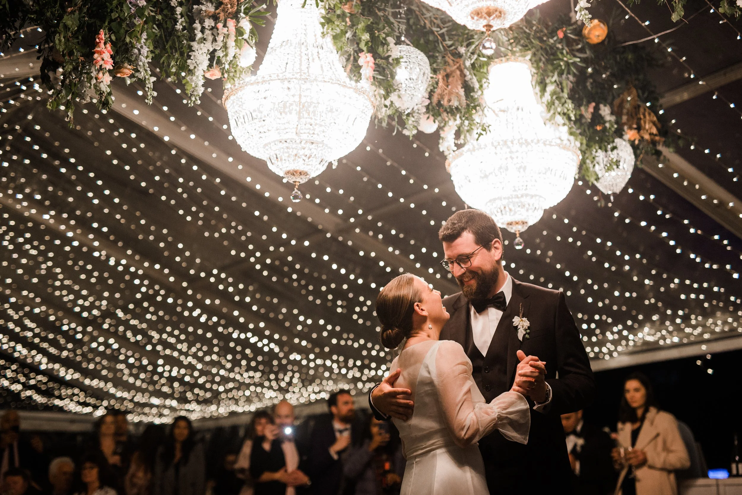 A couple dances at their wedding reception under chandeliers and string lights, surrounded by guests.