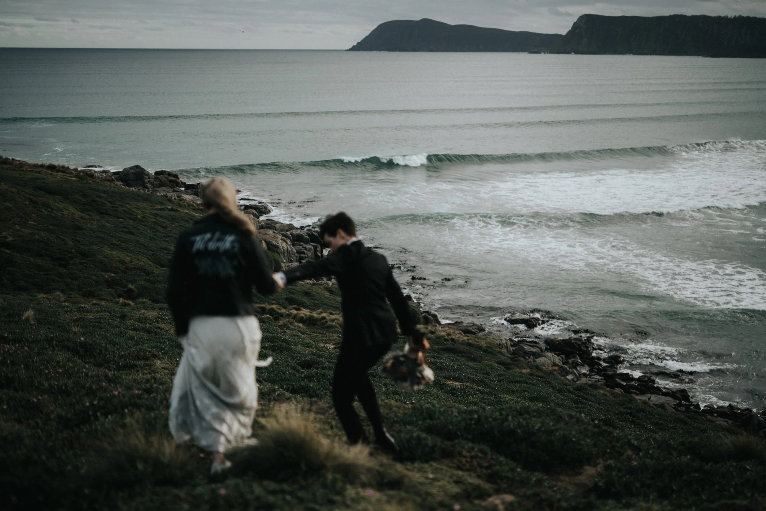 Two people walking along a grassy shoreline of Bruny Island, Tasmania, with waves, with an ocean and cliffs of Bruny Island in the background.