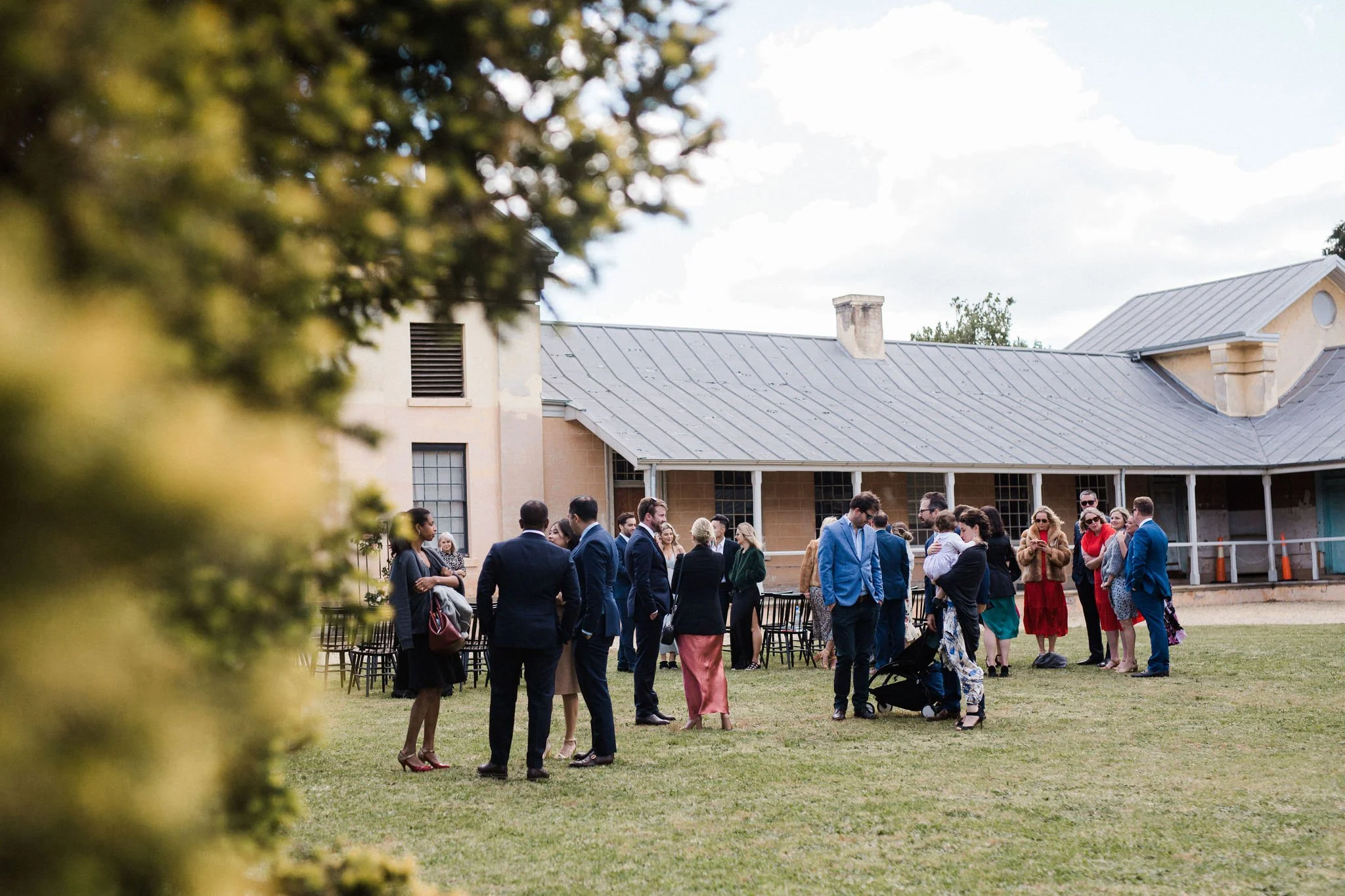 Group of people in formal attire socializing outdoors on a grassy lawn in front of a large building with a metal roof.