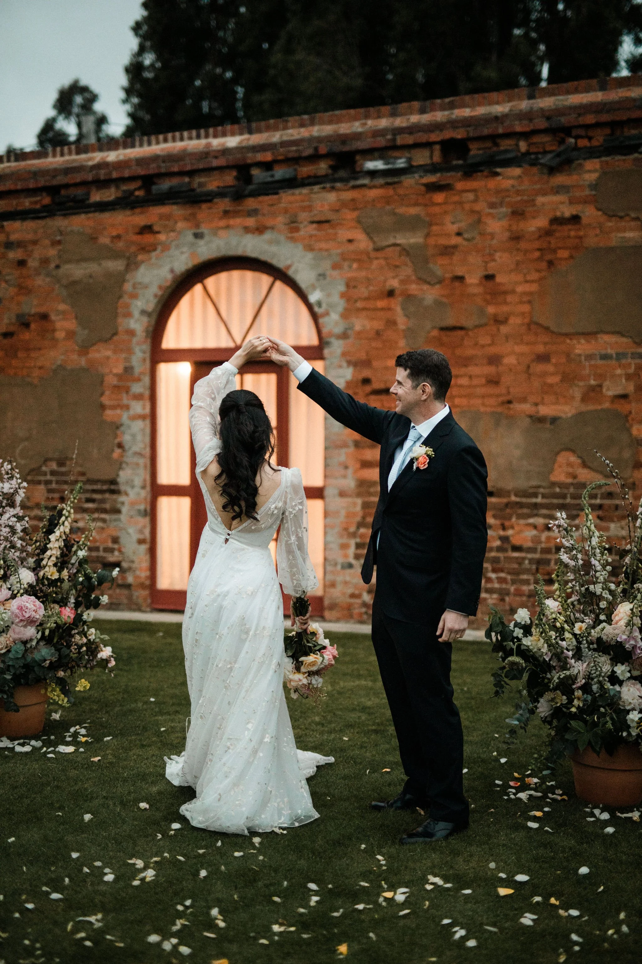 A bride and groom are dancing outdoors during their wedding, with the groom twirling the bride. They are surrounded by flower arrangements, and there's a brick building with illuminated windows in the background.