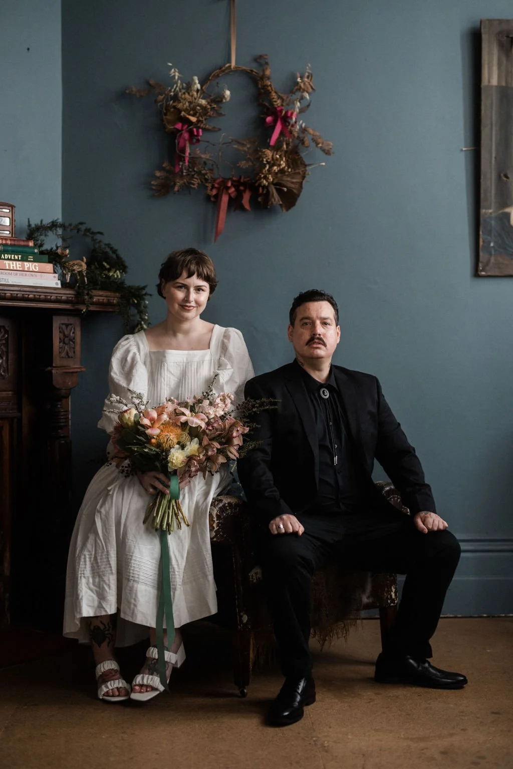 A woman in a white dress holding a bouquet of flowers sitting next to a man in a black suit, both in a vintage room with a dark blue wall and a decorative wreath hanging above them.