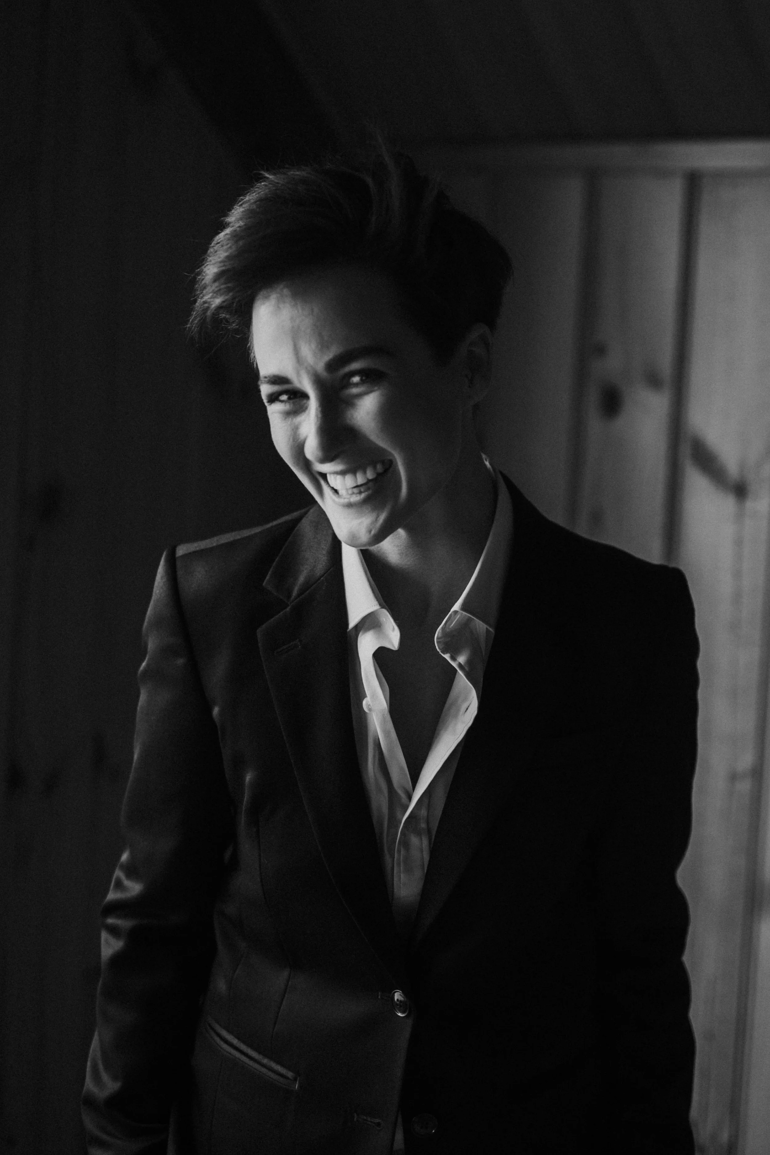 Black and white photograph of a woman in a suit, smiling and showing her teeth, with short hair, against a wooden wall background.