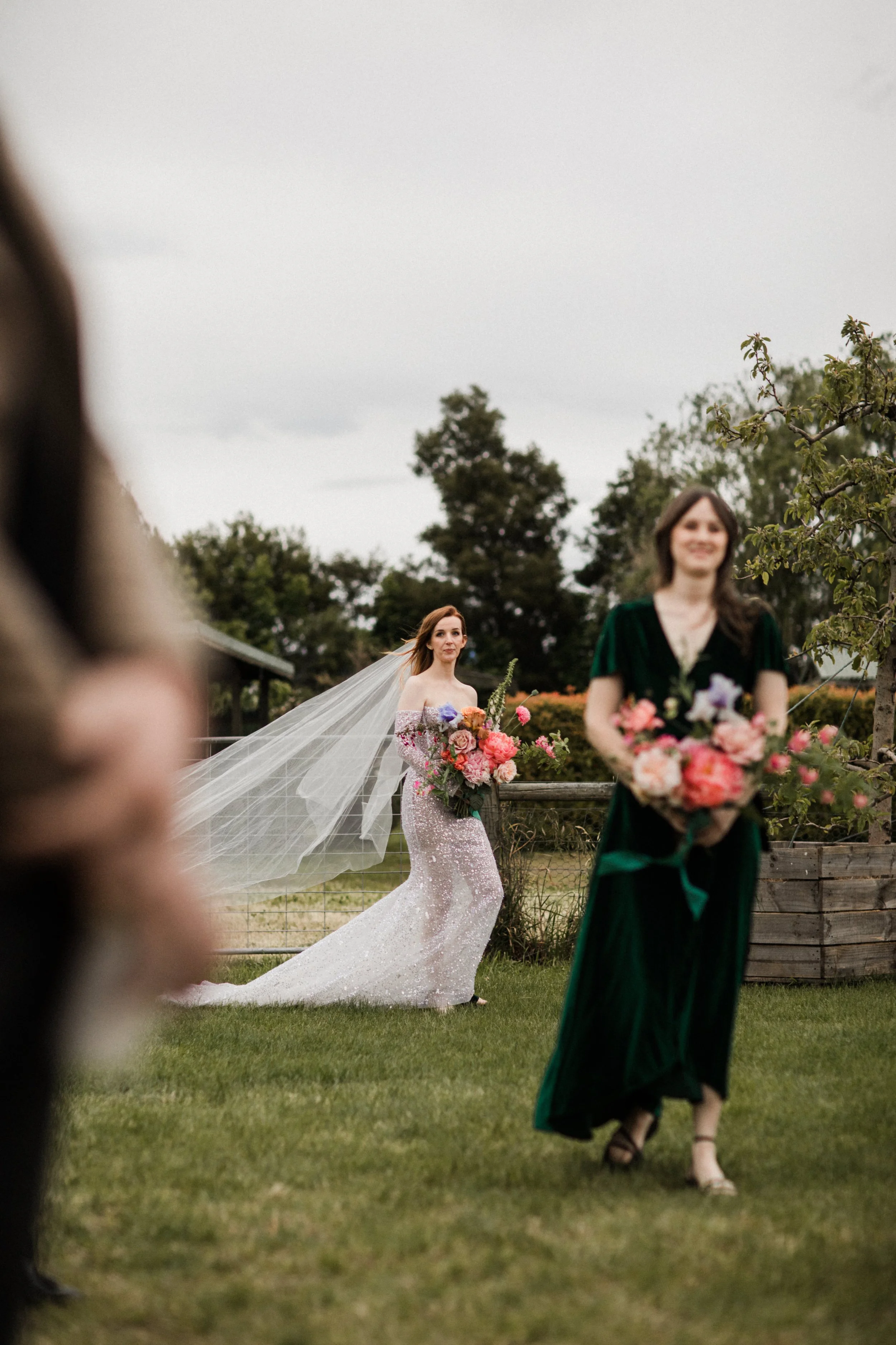 A bride in a strapless wedding gown with a long train and veil holds a bouquet of pink and purple flowers, standing outdoors in a garden. A woman in a green dress with a floral bouquet walks away from her, while others are partially visible in the fo