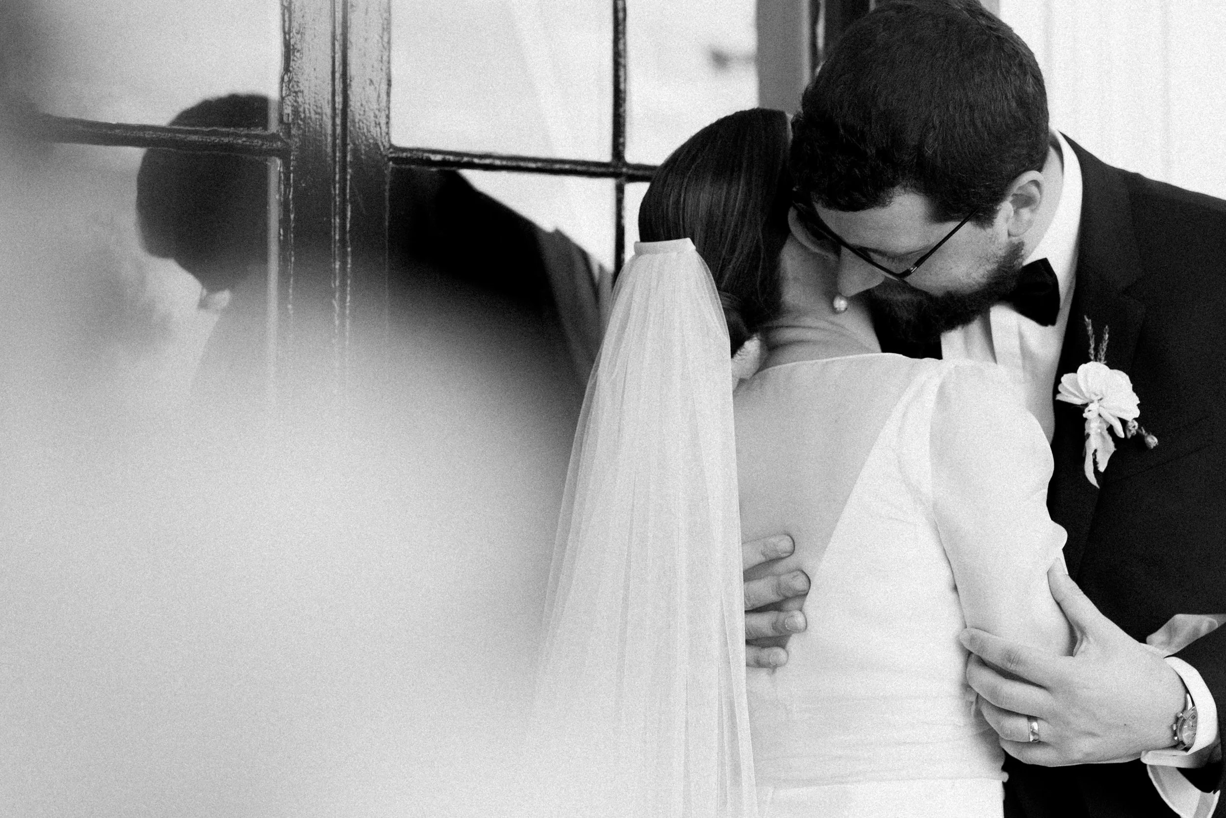 A black-and-white photo of a couple kissing, with the woman wearing a wedding dress and veil, and the man in a suit with glasses and a bow tie, at their wedding.