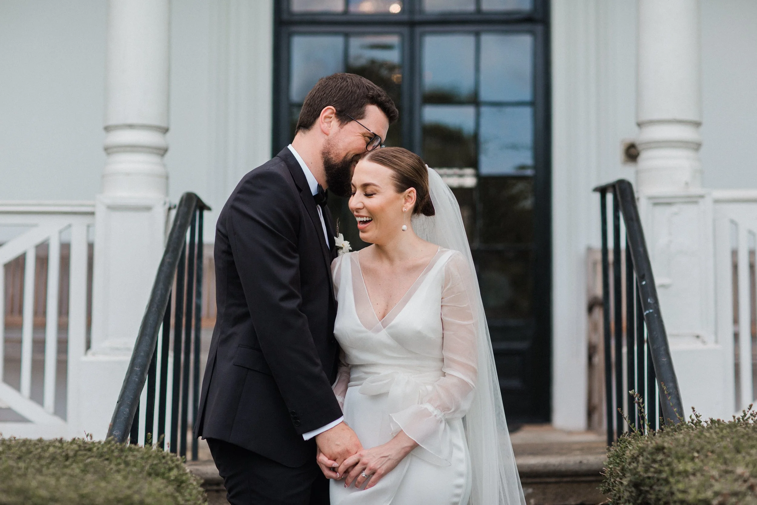 A newlywed couple stands on the steps of a white house, smiling and laughing. The groom wears a black tuxedo with glasses, and the bride wears a white wedding dress with sheer sleeves and a veil.