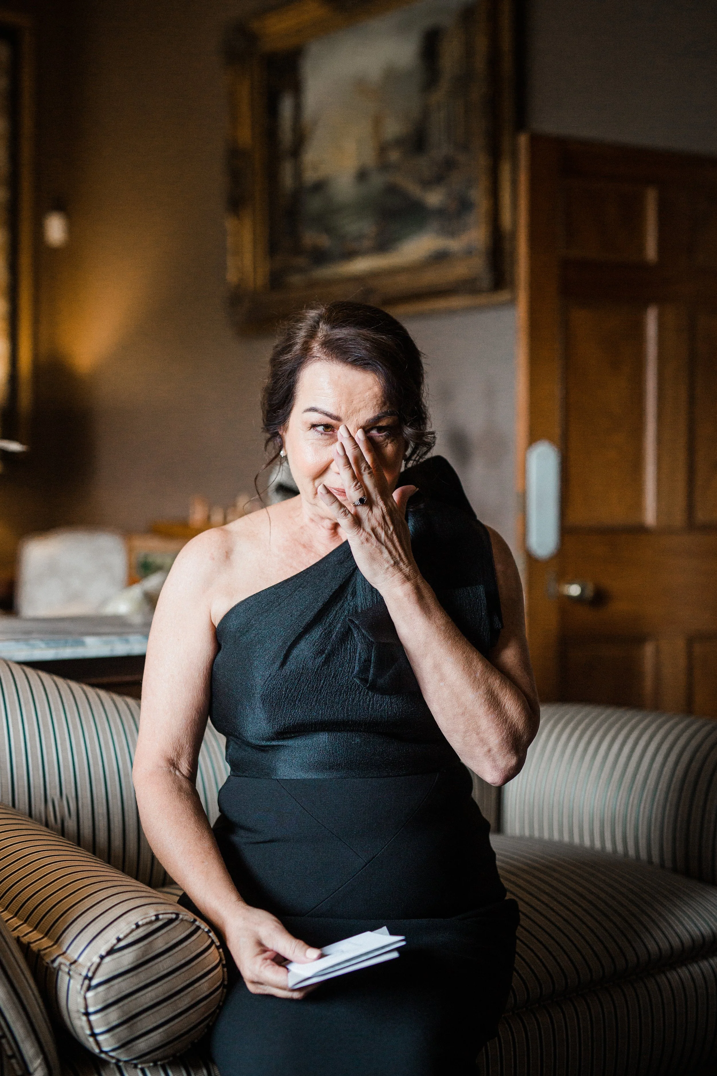 A woman in a black dress sitting on a striped sofa in a room, holding a small stack of papers, with her hand partially covering her face, and looking directly at the camera.