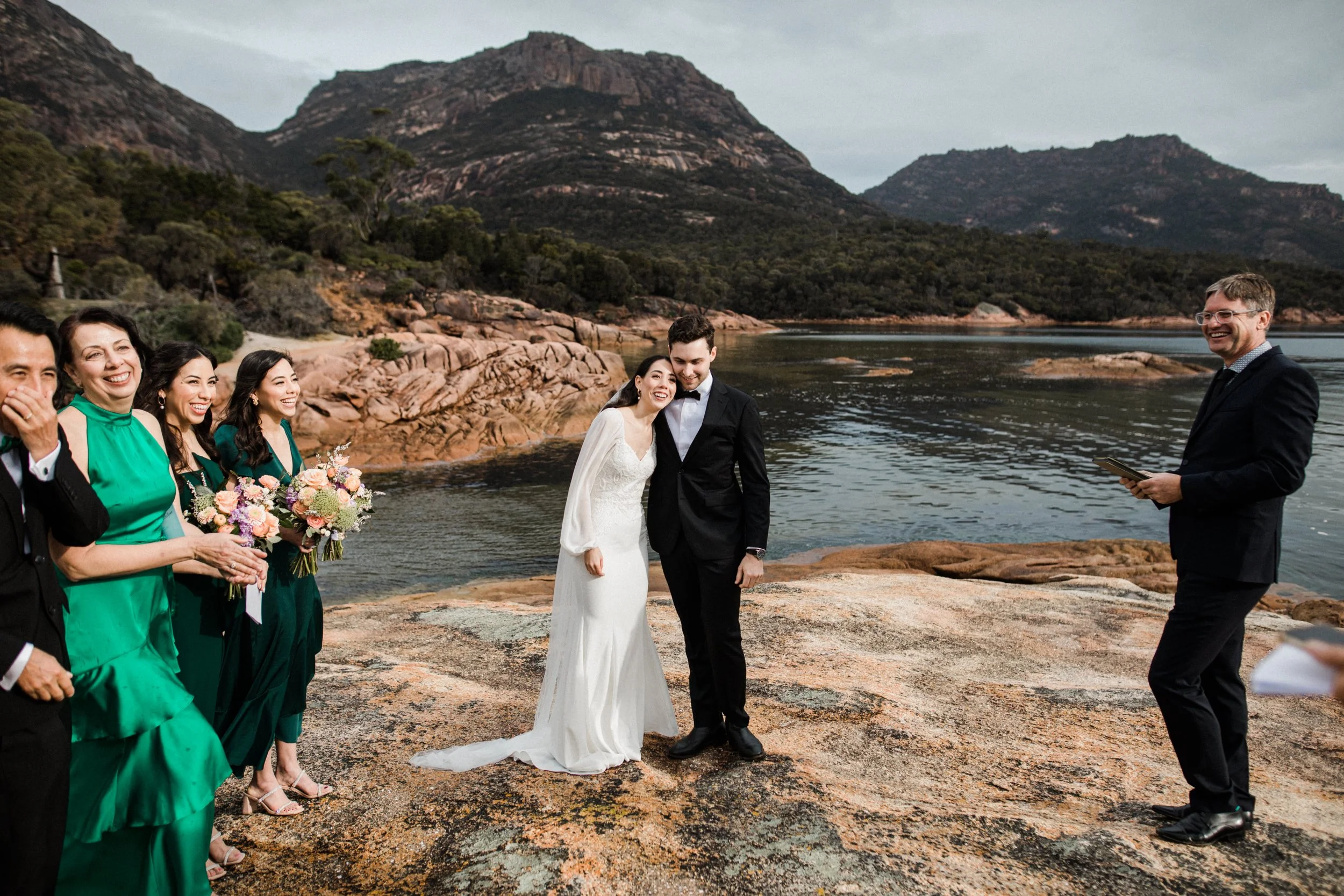 A wedding ceremony taking place outdoors on a rocky waterfront with mountains and water in the background. The bride and groom are standing close together, smiling, with the bride in a white dress and the groom in a black suit. Several bridesmaids an