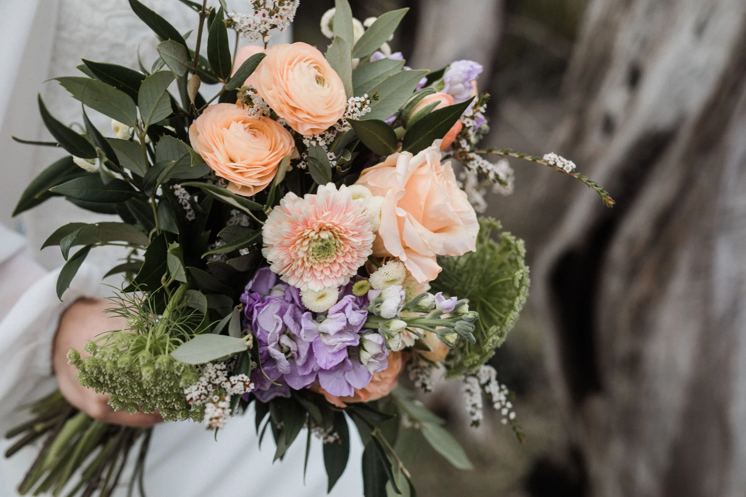 A bouquet of various flowers including peach roses, purple hydrangeas, pink and white ranunculus, and other small white and purple flowers, surrounded by green leaves and stems.
