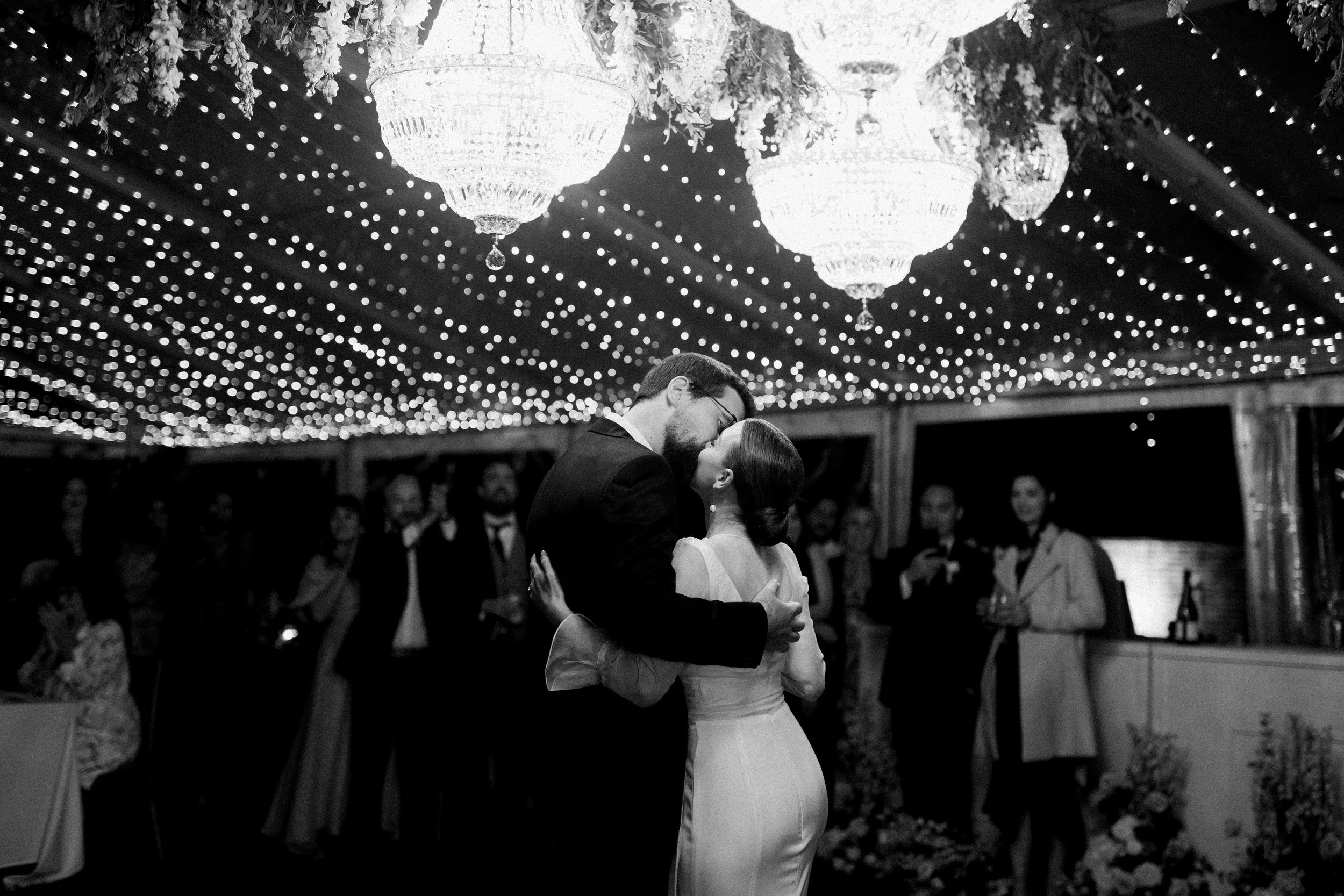 Black and white photo of a couple dancing closely at their wedding reception, under hanging chandeliers and string lights, with guests watching in the background.
