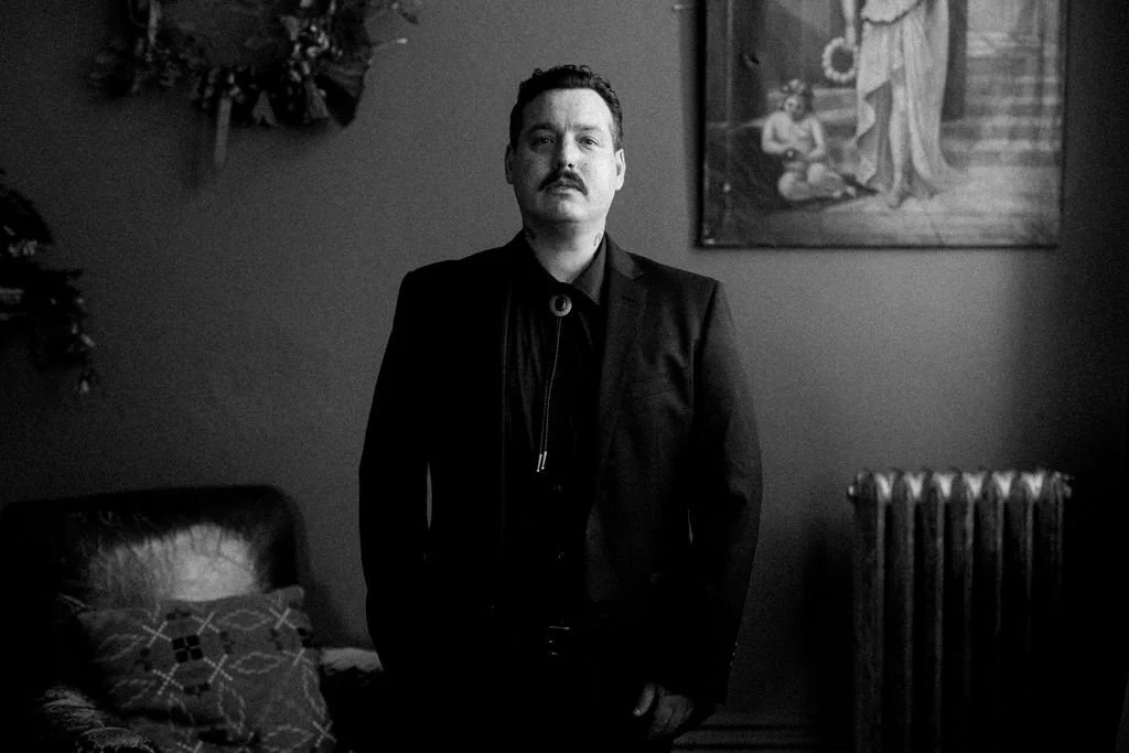 A man with a mustache in a black suit standing in a room with a painting on the wall, a radiator, and a sofa with patterned pillows.