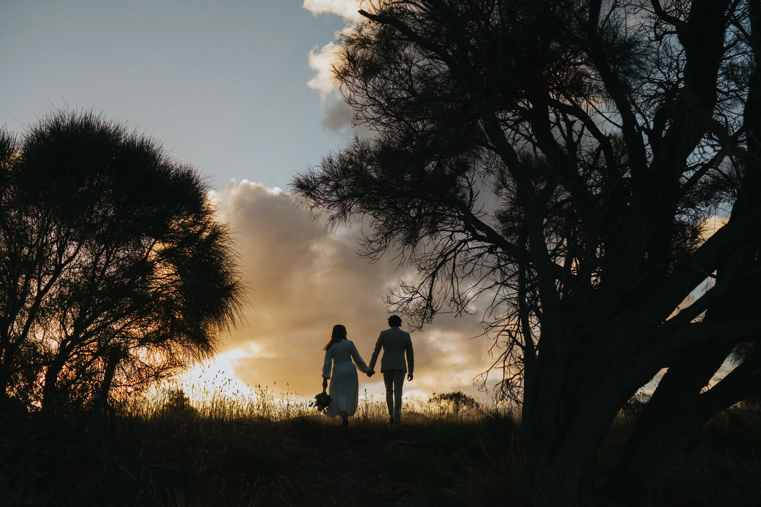 A couple walking hand in hand through a grassy field at sunset, silhouetted against the sky with trees on either side.