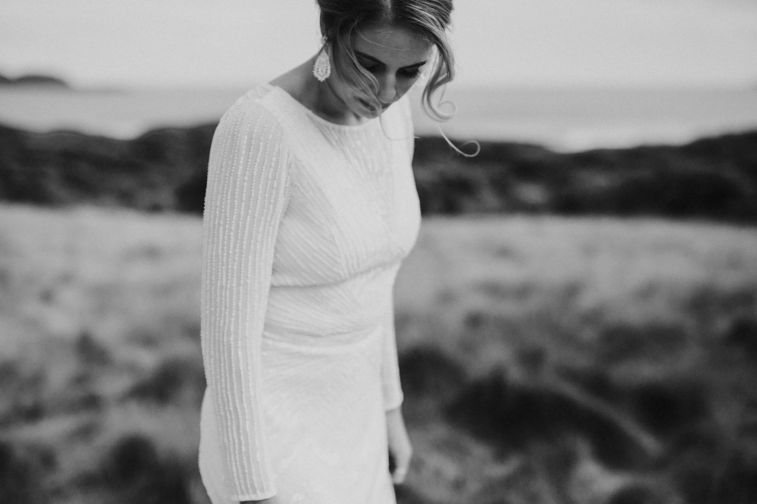 Black and white photo of a woman in a long dress with textured patterns, standing outdoors with rocks in the background, looking down.