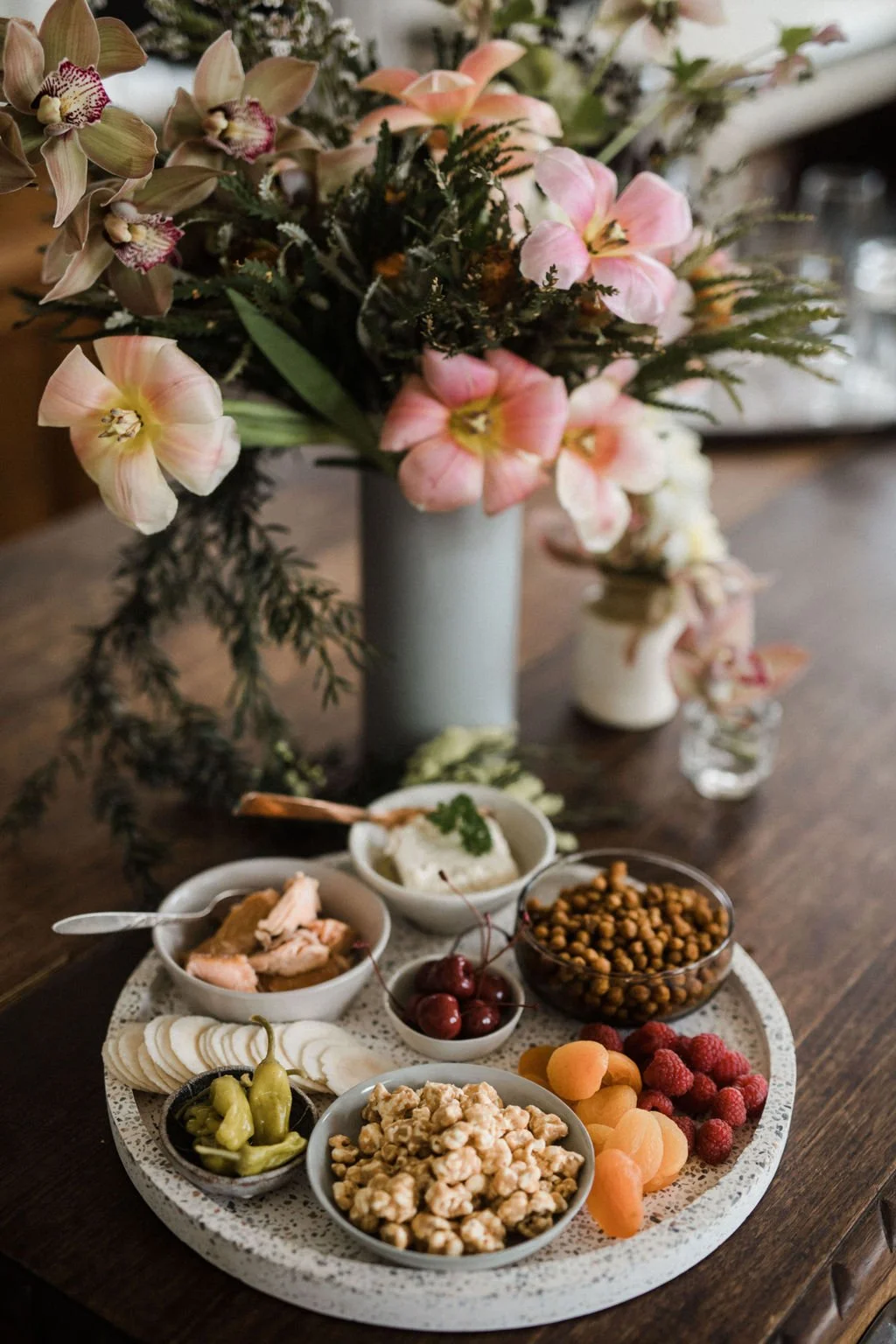 A flower arrangement with pink and beige orchids and greenery in a tall, pale gray vase. In front, a round tray holds bowls of assorted snacks, including cherries, raspberries, apricots, popcorn, pickled peppers, and other small bites, on a wooden ta
