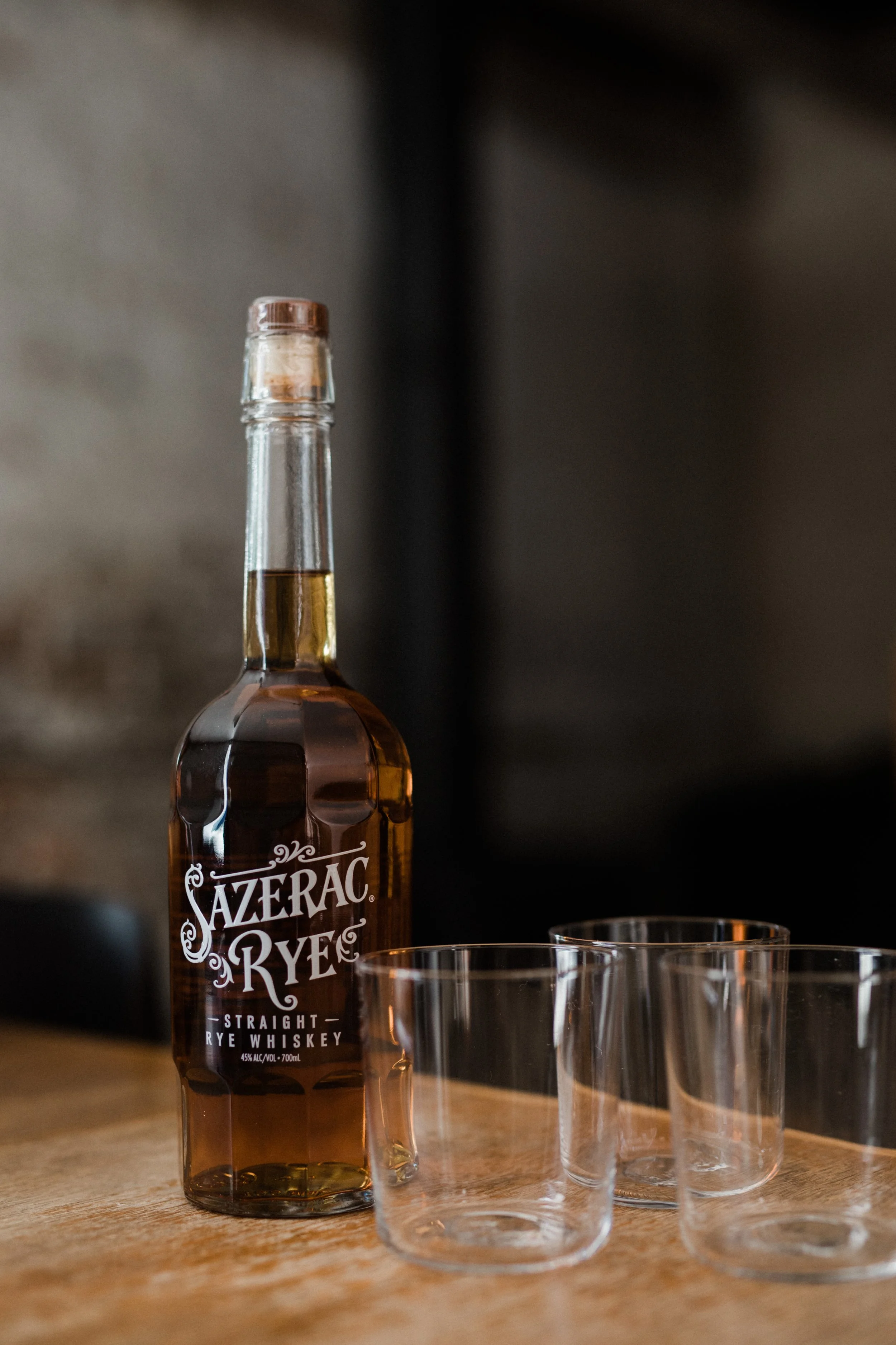 A bottle of Sazerac Rye whiskey and three empty glass tumblers on a wooden surface.