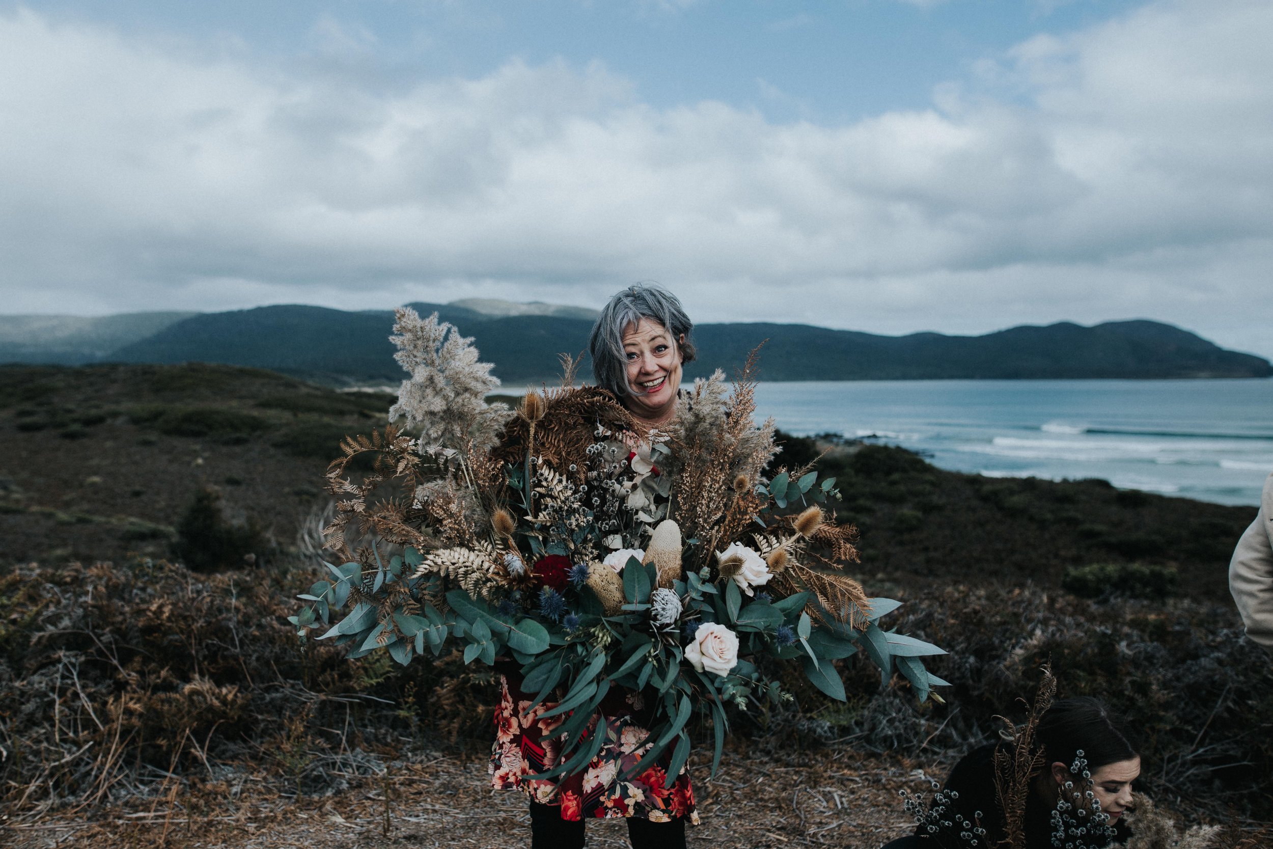 A smiling woman holding a large bouquet of flowers and dried plants on a coastal landscape with mountains and ocean in the background.