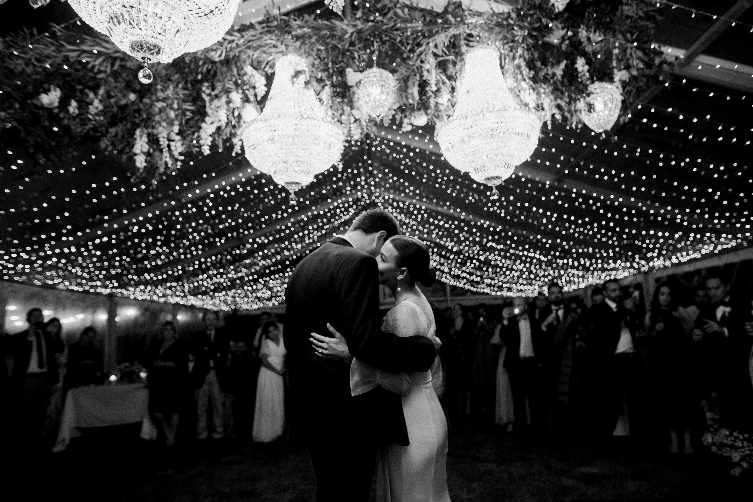 A black and white photo of a couple dancing closely under decorative chandeliers and string lights at a wedding reception, with guests watching in the background.