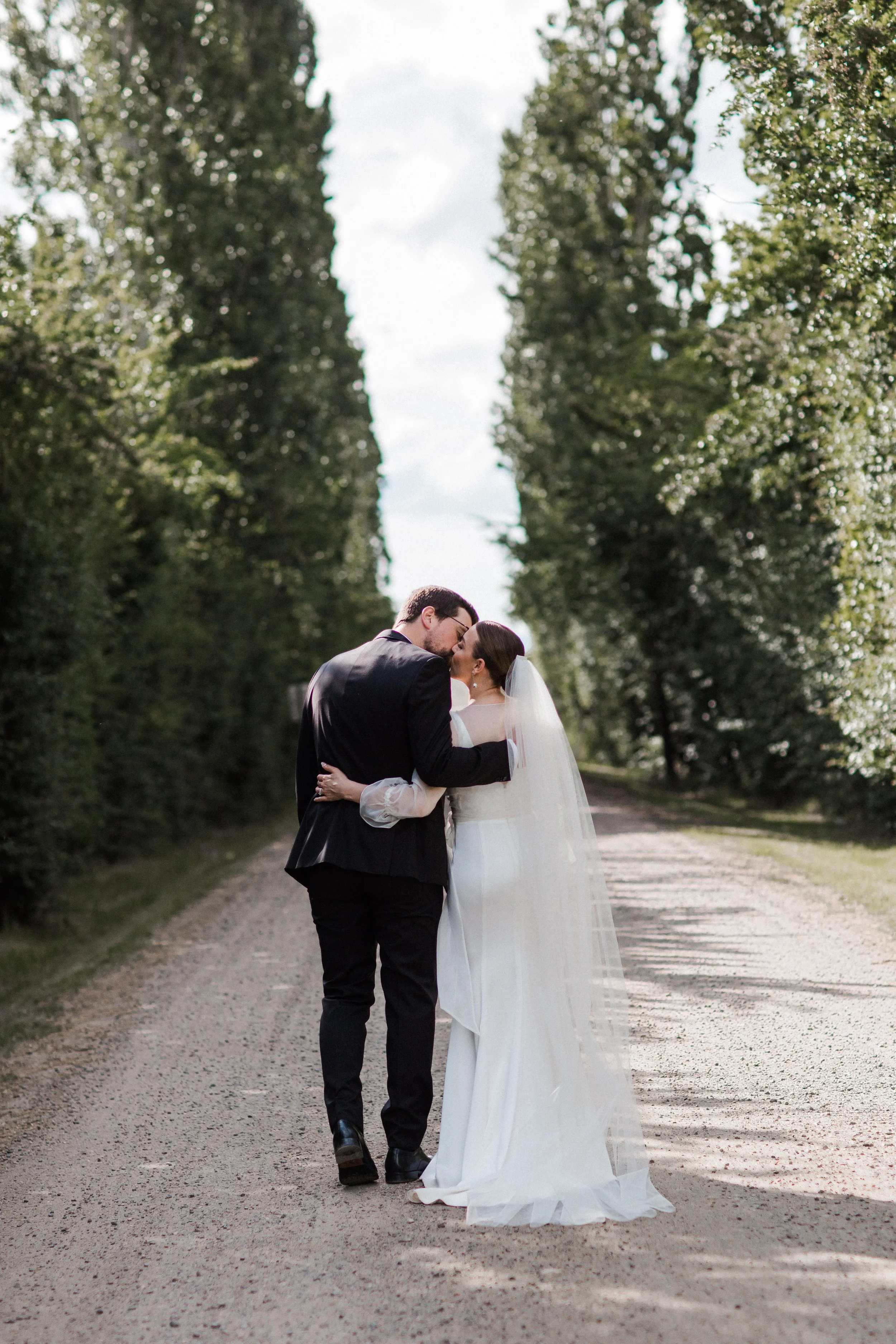 A newlywed couple shares a kiss on a tree-lined dirt road.