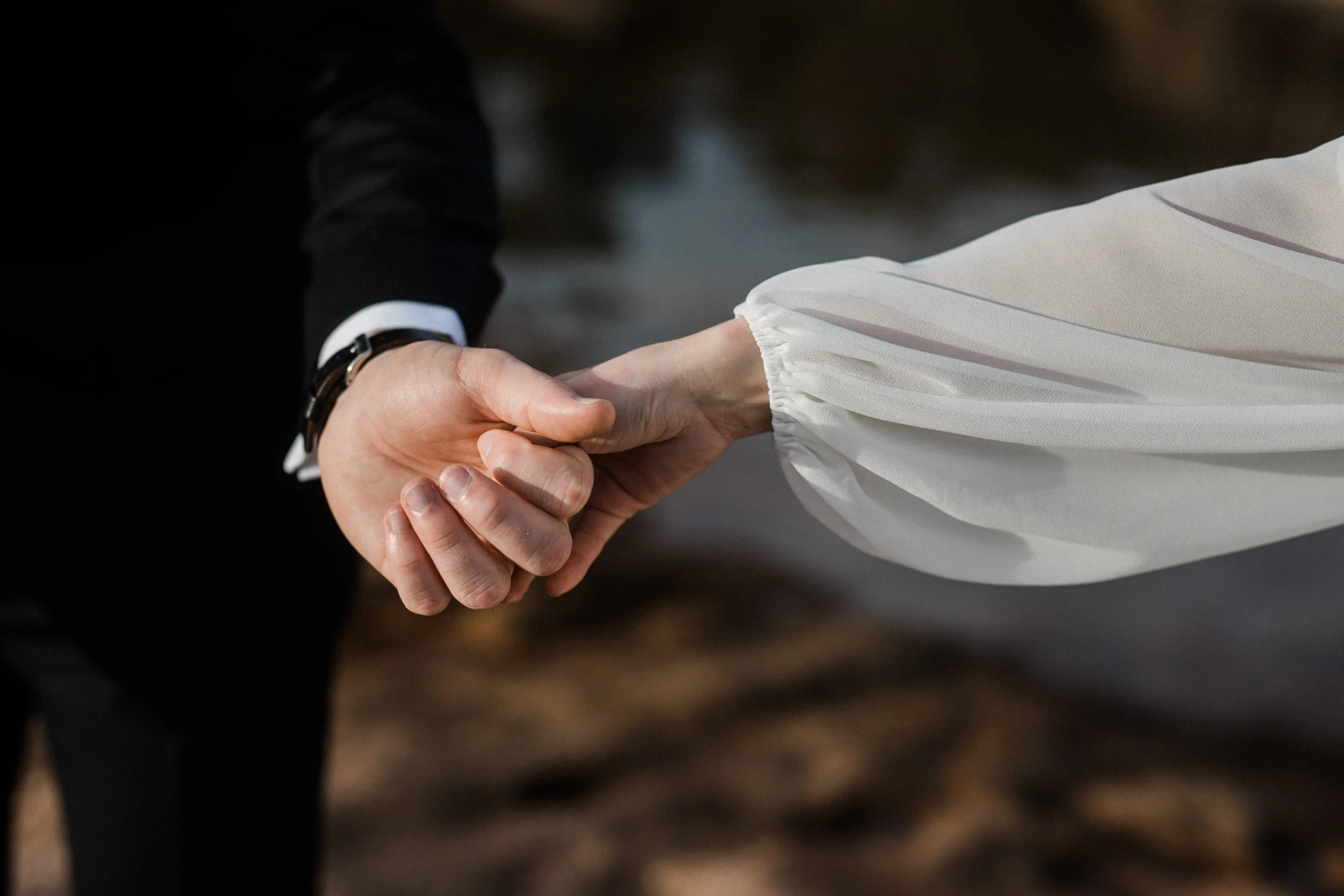 Close-up of a groom and bride holding hands during a wedding, with a blurred outdoor background.