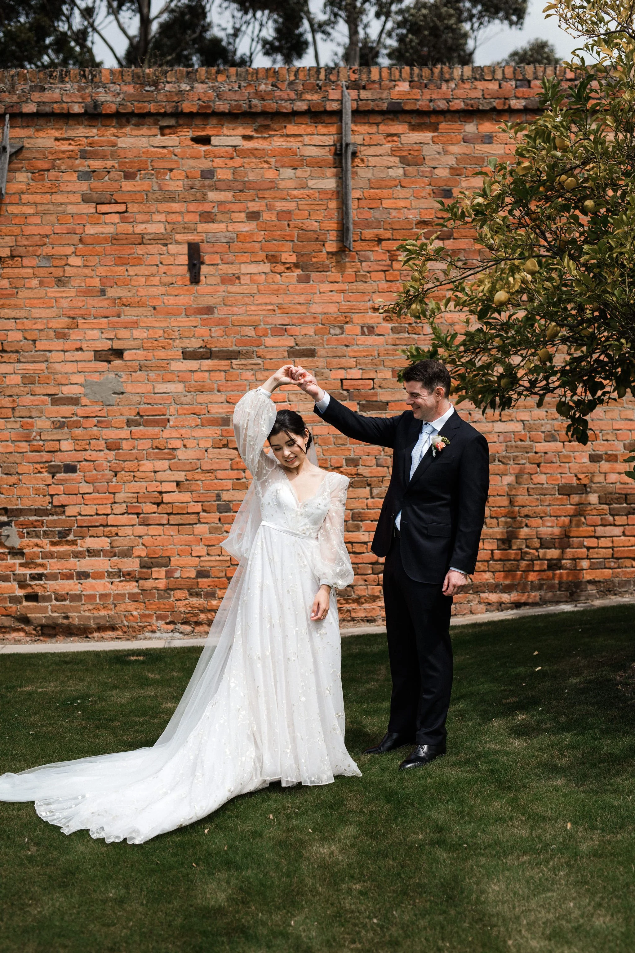 A bride and groom dancing outdoors in front of a brick wall, with the groom twirling the bride.