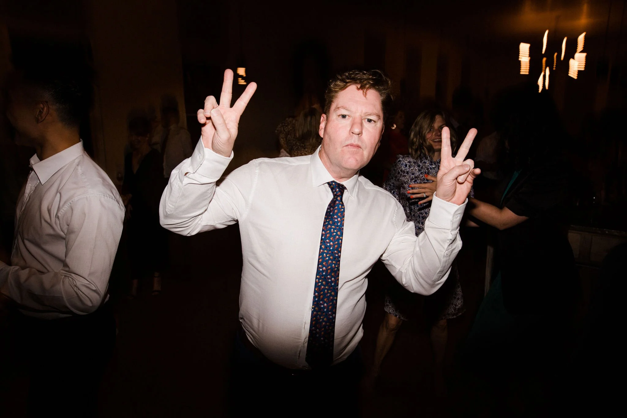 Man making a peace sign with both hands at a social event, with other people dancing and talking in the background.