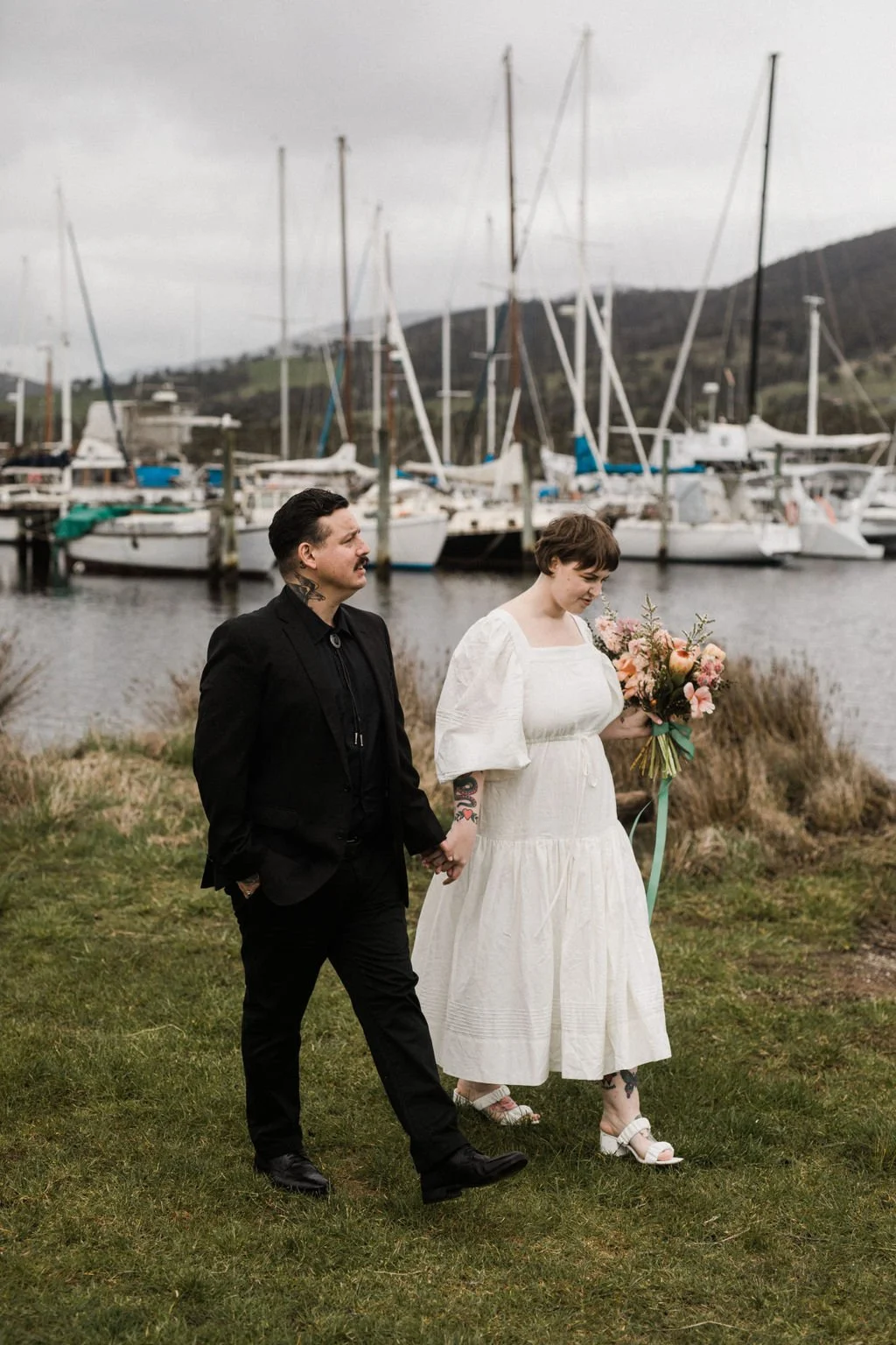 A couple holding hands walks by a marina with sailboats and yachts in the background. The woman is wearing a white dress and carrying a bouquet of flowers, while the man is dressed in black.