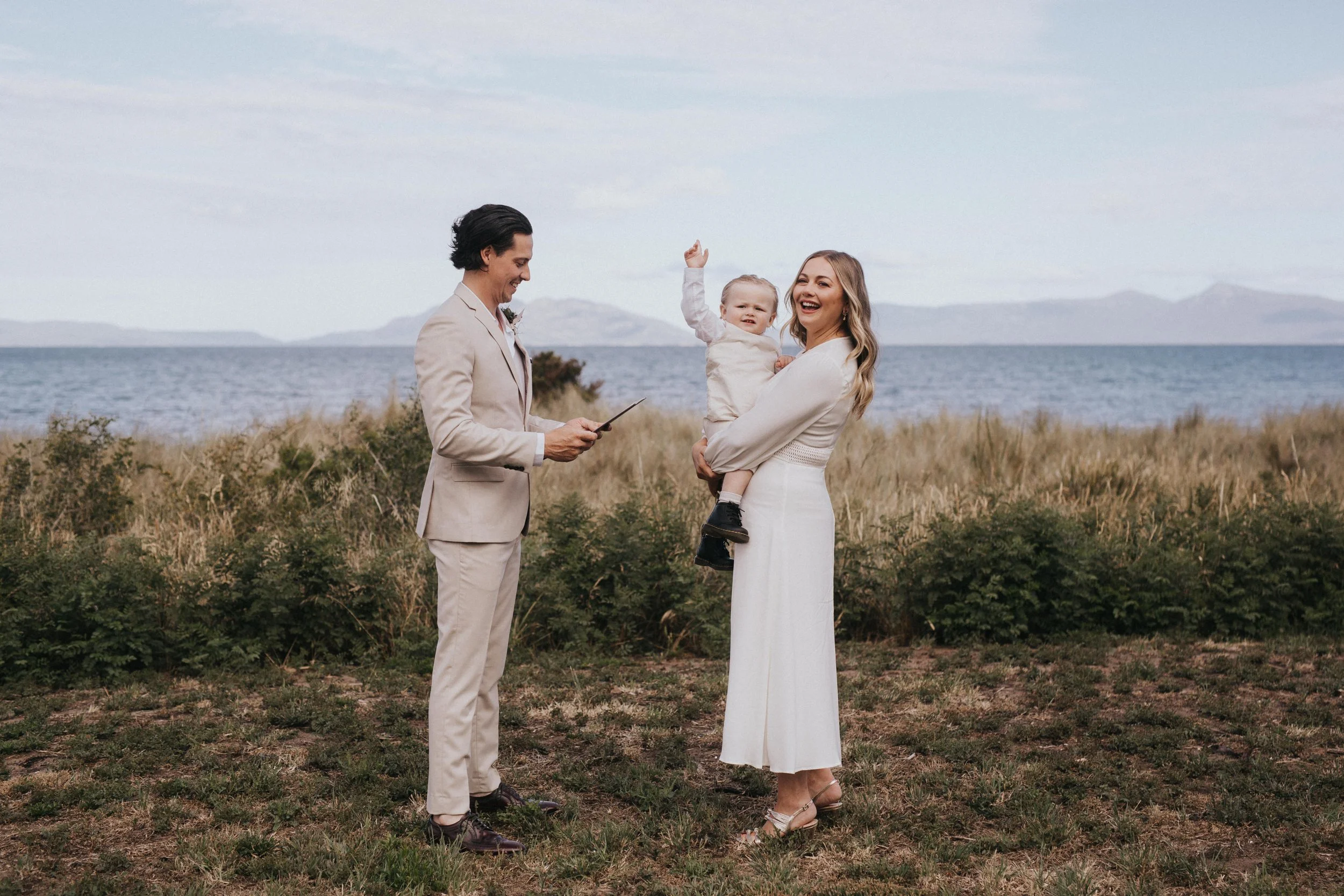 A couple and child standing on a grassy area near the water, with a man in a beige suit reading vows or a speech, a woman in a white dress holding a smiling child raising a hand, and a lake with mountains in the background.
