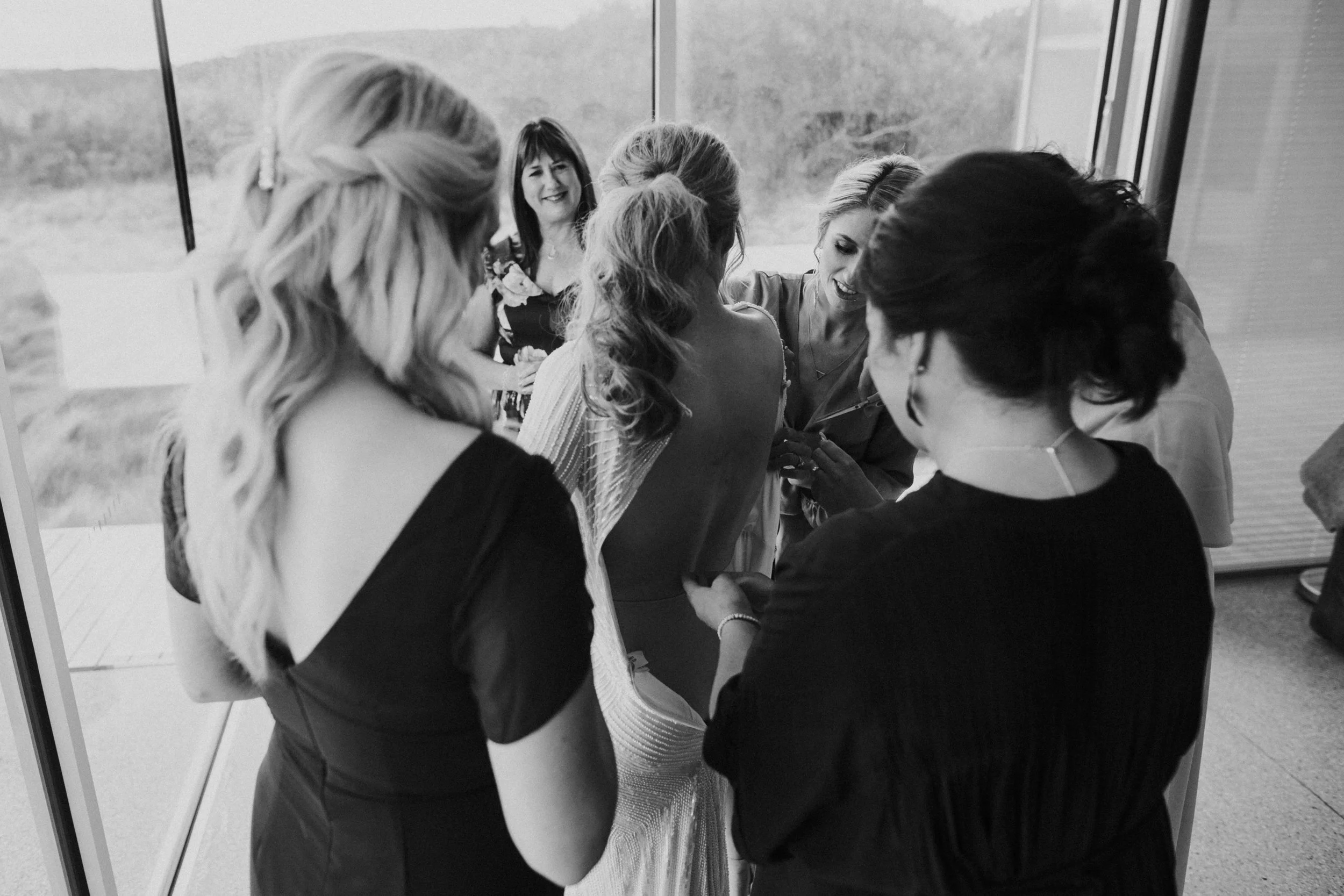 A group of women gathered in a room, some smiling and laughing, as they a prepare a woman for her wedding, possibly for a bridesmaid or wedding dress fitting.